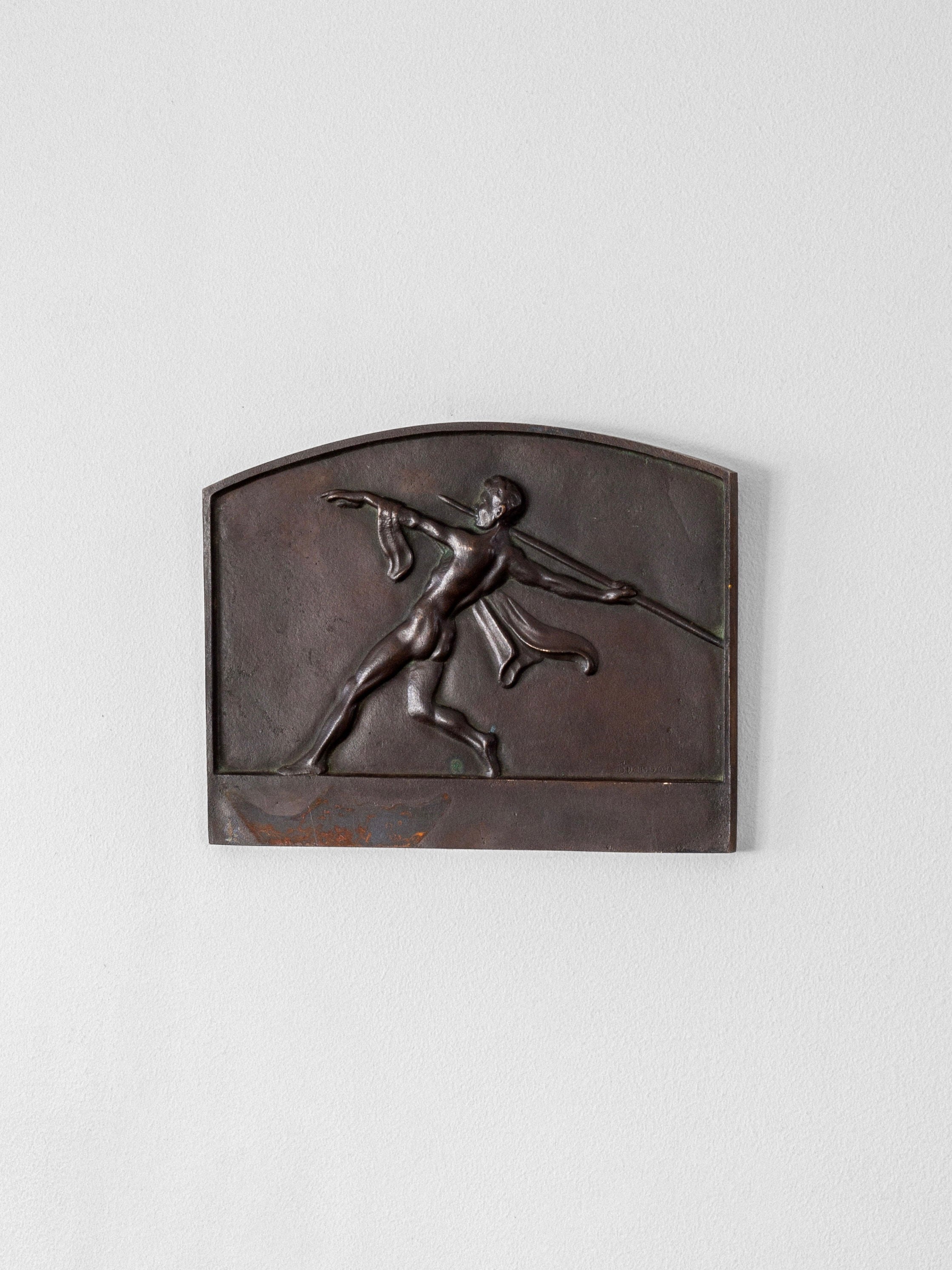 The Spigel Art Deco Bronze Wall Relief showcases a nude male figure with a spear and cloth, dynamically posed against a rectangular background with an arched top. Mounted on a light gray wall, this dark-toned piece evokes classical elegance.