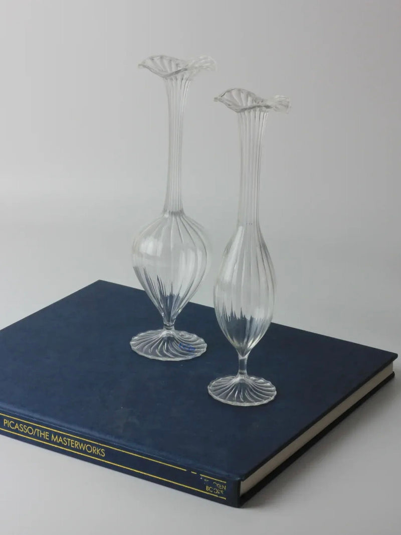 Two vintage Murano candle holders from Boga Avante Shop, with elegant narrow necks and flared tops, are displayed on a large blue book titled "Picasso/The Masterworks" against a light backdrop. This arrangement offers a minimalist, artistic display embodying classic timeless style.