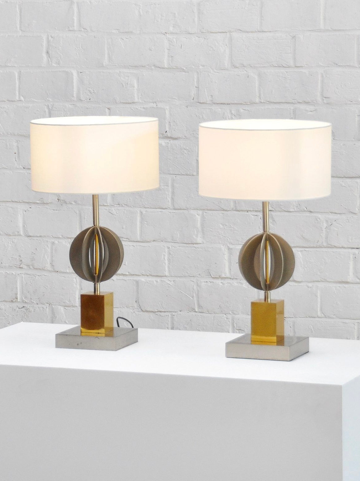 The Pair of Italian Geometric Table Lamps 70's by Introverso features two lamps with white cylindrical shades and brass and chrome finishes, highlighted by a central spherical design and square bases. They're showcased on a white platform against a white brick wall.