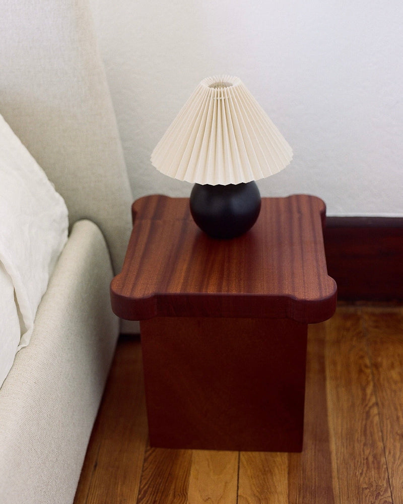 A small, dark wooden bedside table with a wavy edge from MUHLY, known as the Bijou Mini Table, sits on a wooden floor beside a light-colored bed. A black lamp with a pleated white shade is placed on the table. The white wall behind and minimalist design complete the serene space.