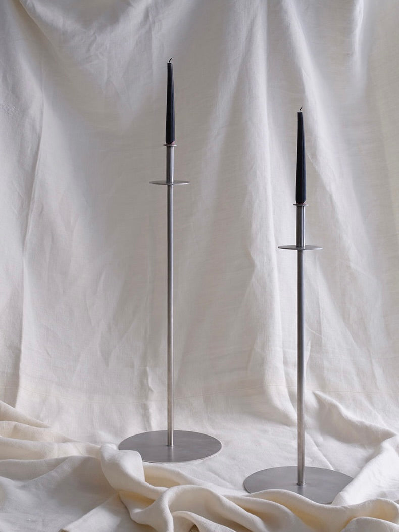 Two tall FEBRERO SHOP Fusta Candle Holders made of brushed stainless steel, each holding a black taper candle, sit on a textured light-colored fabric backdrop, creating a minimalist and elegant aesthetic.