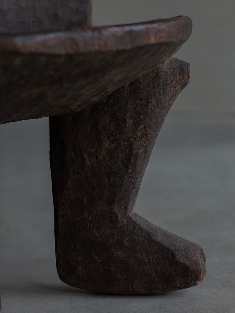 A close-up of the African Stool by Bicci de' Medici features a carved wooden leg resembling a boot, crafted by African artisans. The wood has a dark, textured finish that reflects cultural heritage, set against a smooth, neutral-colored background.