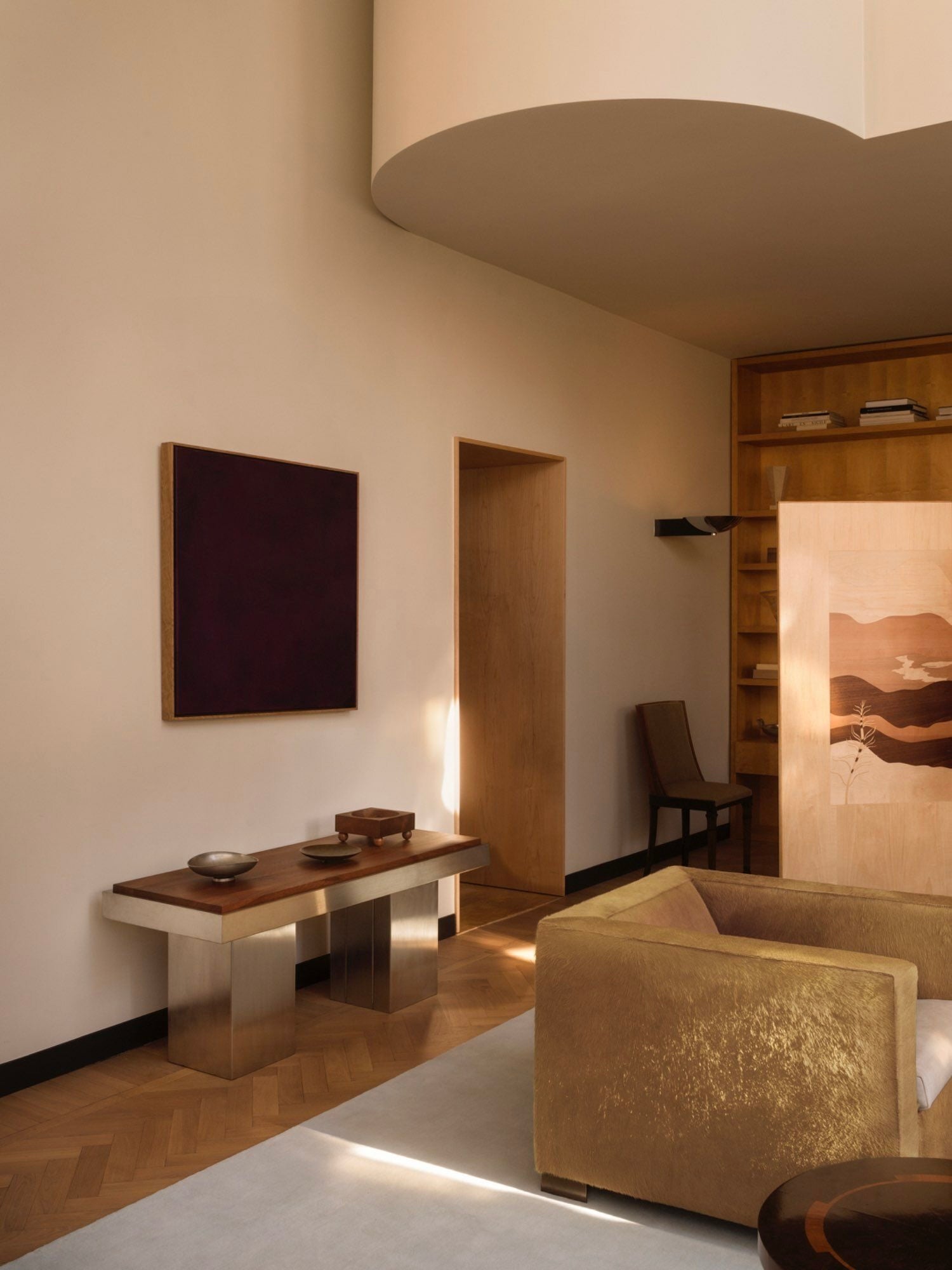 A modern living room with minimalist design elements inspired by the Bauhaus movement. It showcases a gold-toned couch, the Studio Vraco "Console Table 02," which features a wooden surface and brushed stainless steel frame, dark rectangular wall art, and light wood shelving. The space is well-lit with neutral tones and parquet flooring.