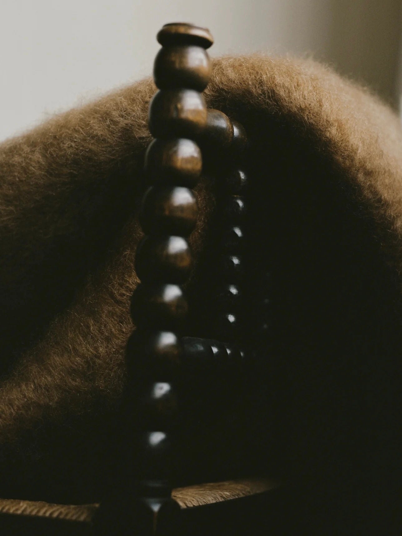 A close-up of a dark wooden chair with a distinctive spindle design features "Arbor Sable" Throw by The Wren draped over the back, adding warmth to the scene. Soft, natural lighting creates a cozy, inviting atmosphere.