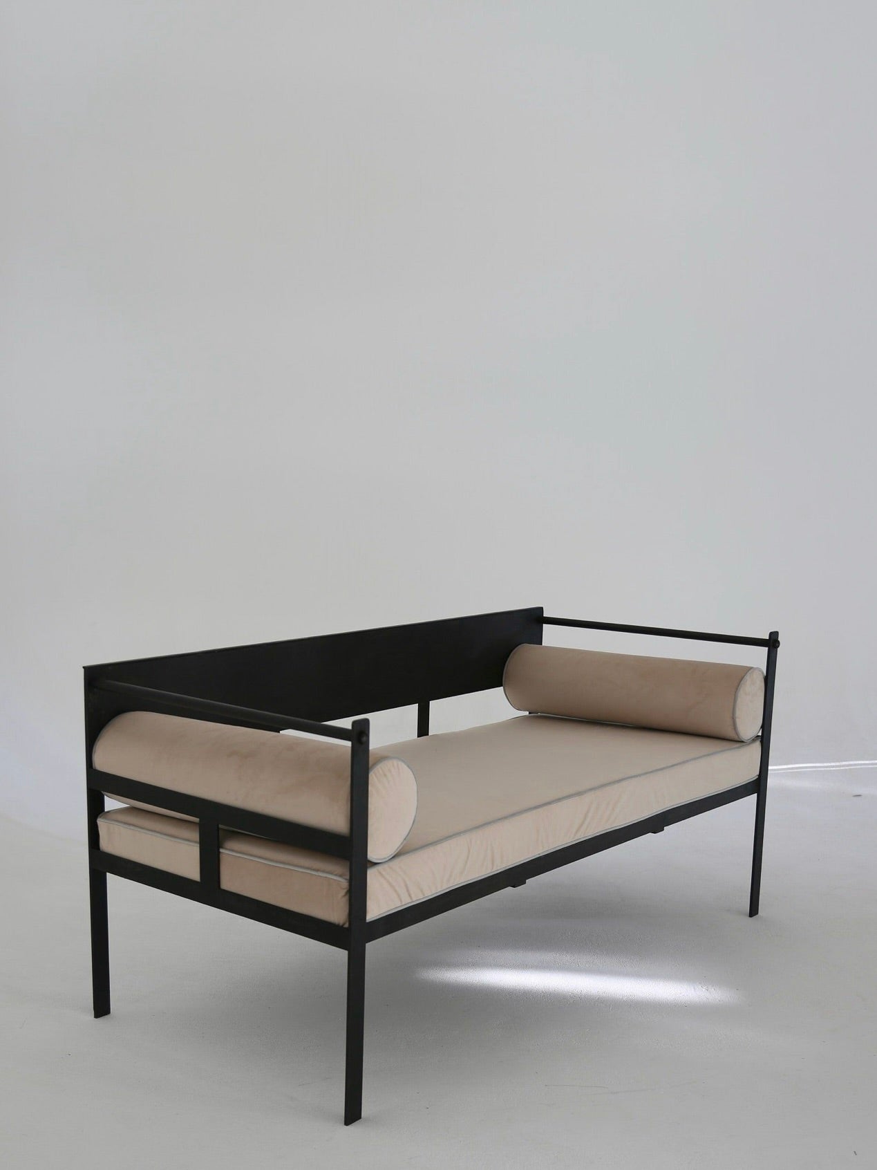 The Ancien et Jolie Modernist Lounge Set features a minimalist black metal-framed daybed, inspired by Mathieu Mategot, with beige cushions and cylindrical bolster pillows on each side, showcased on a plain white background.