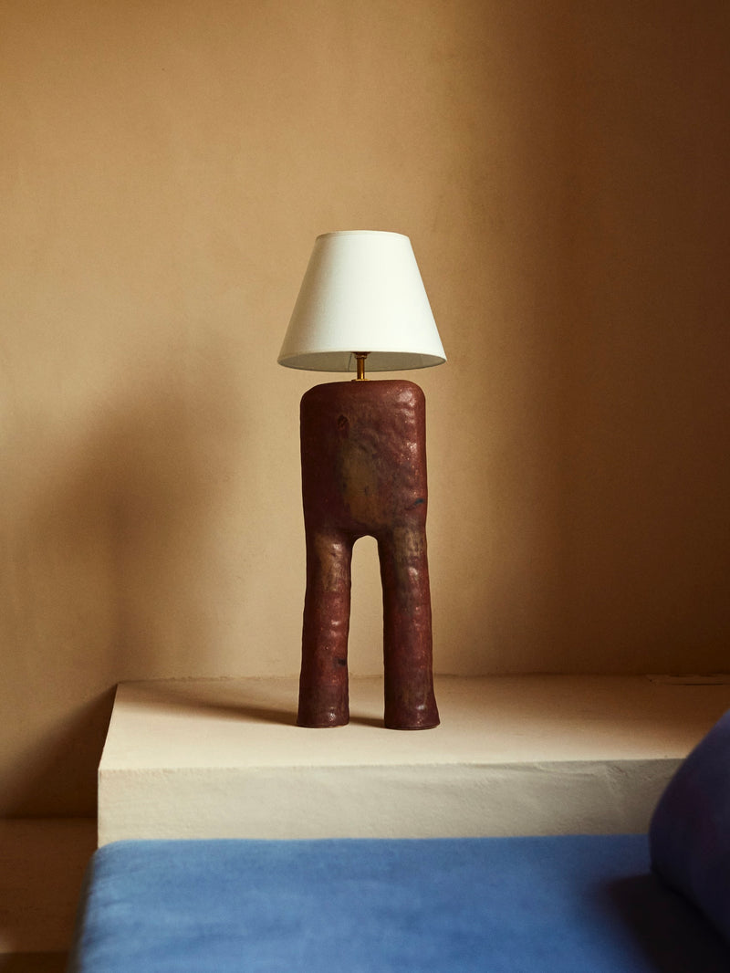 The LIV Table Lamp by marta bonilla features a unique handcrafted design with a sculpted, matte brown glazed base resembling abstract shapes. It stands on a white surface adorned with a simple white conical lampshade, while soft blue fabric appears in the foreground.
