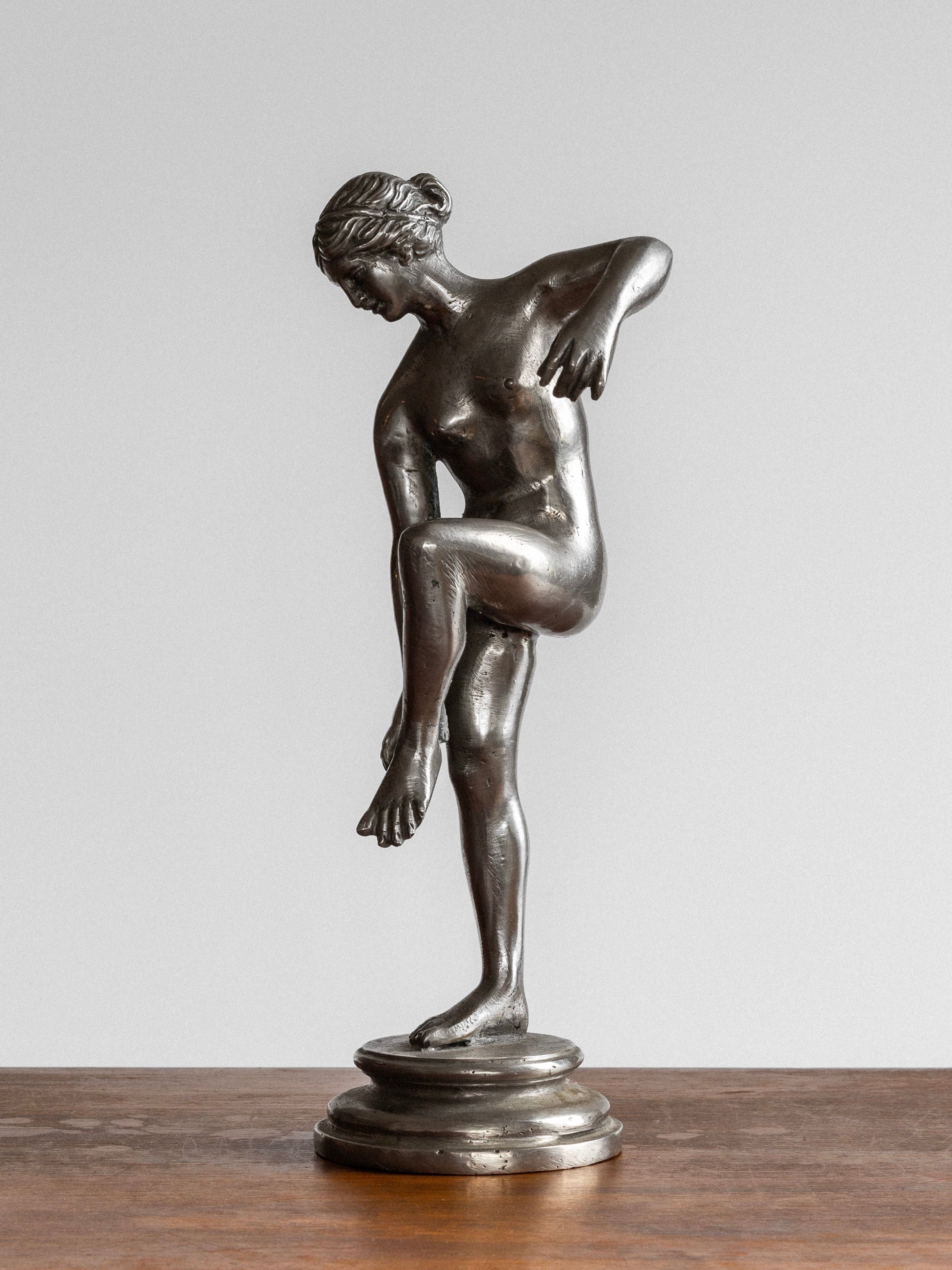 The Spigel Elegant Metal Sculpture from the 1930s features a graceful nude woman balanced on one leg, her right arm extended downward and left leg bent, with her left hand touching her extended leg. This neoclassical design is mounted on a circular pedestal and placed on a wooden surface.