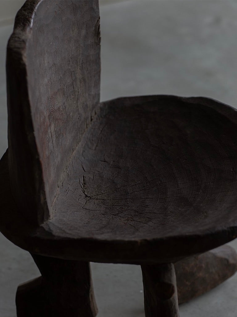 The African Stool by Bicci de' Medici features a rustic wooden design with a textured, curved backrest and seat, highlighting primitive elements. Aged wood with visible grain patterns and cracks showcases the skilled craftsmanship of African artisans on this light-colored surface.