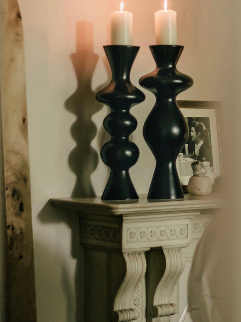 Two ceramic candleholders from Ransom & Dunn, each with a lit candle, sit on an ornate white shelf. Accompanying them are a framed black-and-white photo, a small sculpture of the female form, and a small round object, all casting soft shadows on the wall.