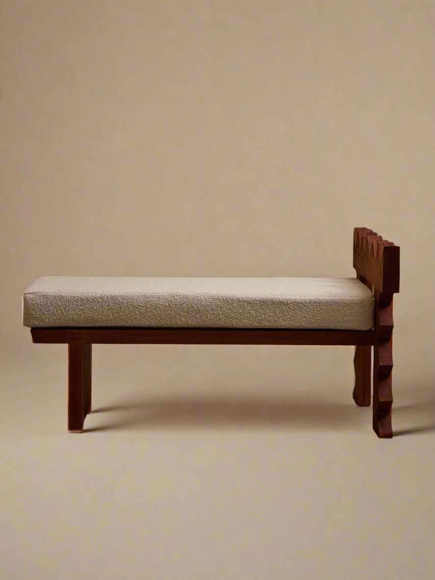 The Jura Bench by Ah Um Design Studio, with its cushioned white seat and sculptural form, sits gracefully against a backdrop of beige curtains. This solid mahogany piece showcases exquisite hand-carved details on the backrest, effortlessly merging minimalist and modern design elements. The floor appears to be made of concrete.