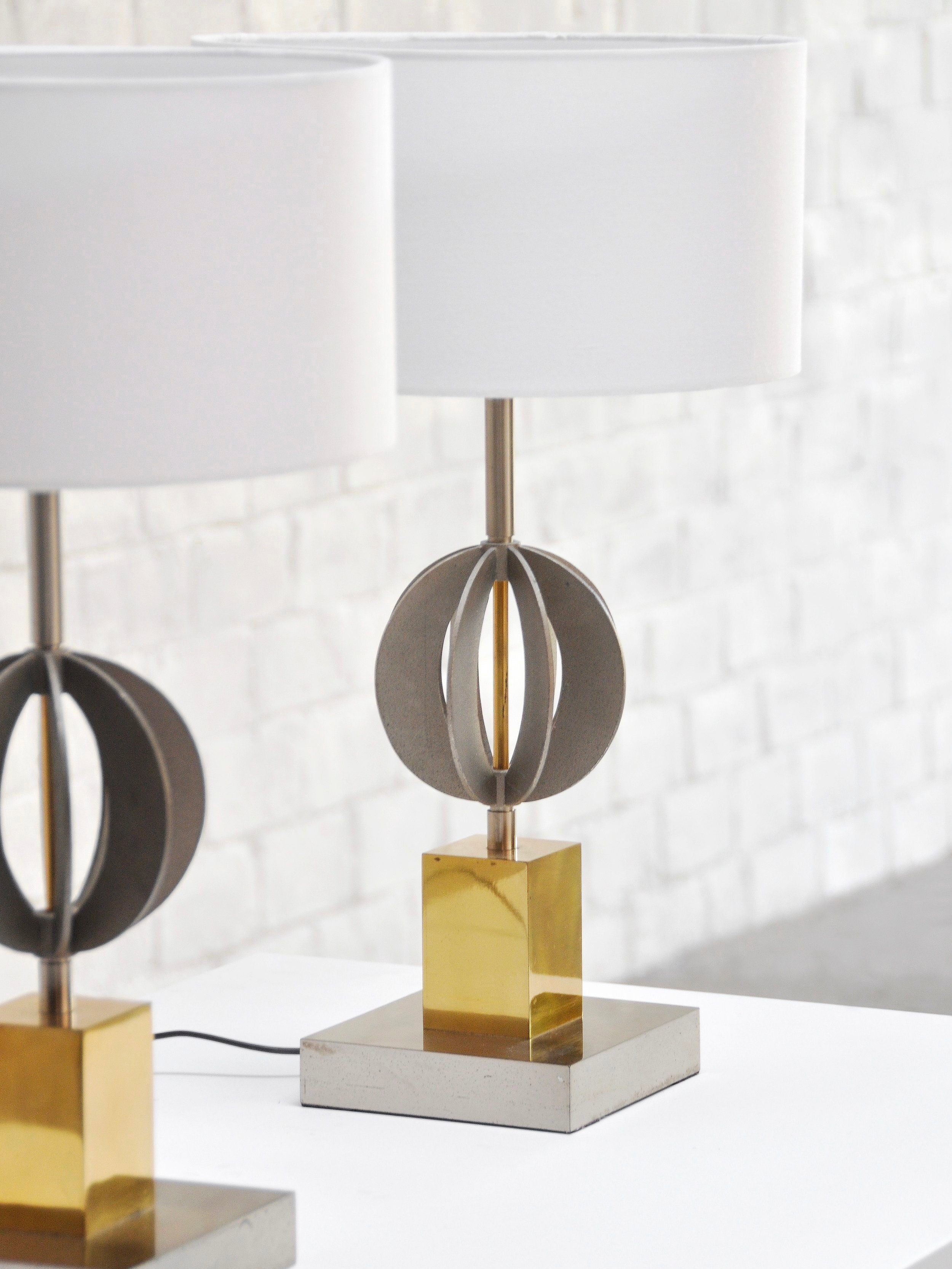 The Pair of Italian Geometric Table Lamps 70's by Introverso boast modern geometric bodies with metal circular designs and chrome and brass accents, topped with white cylindrical shades on gold square bases. They sit on a white table against a sleek white brick wall backdrop.