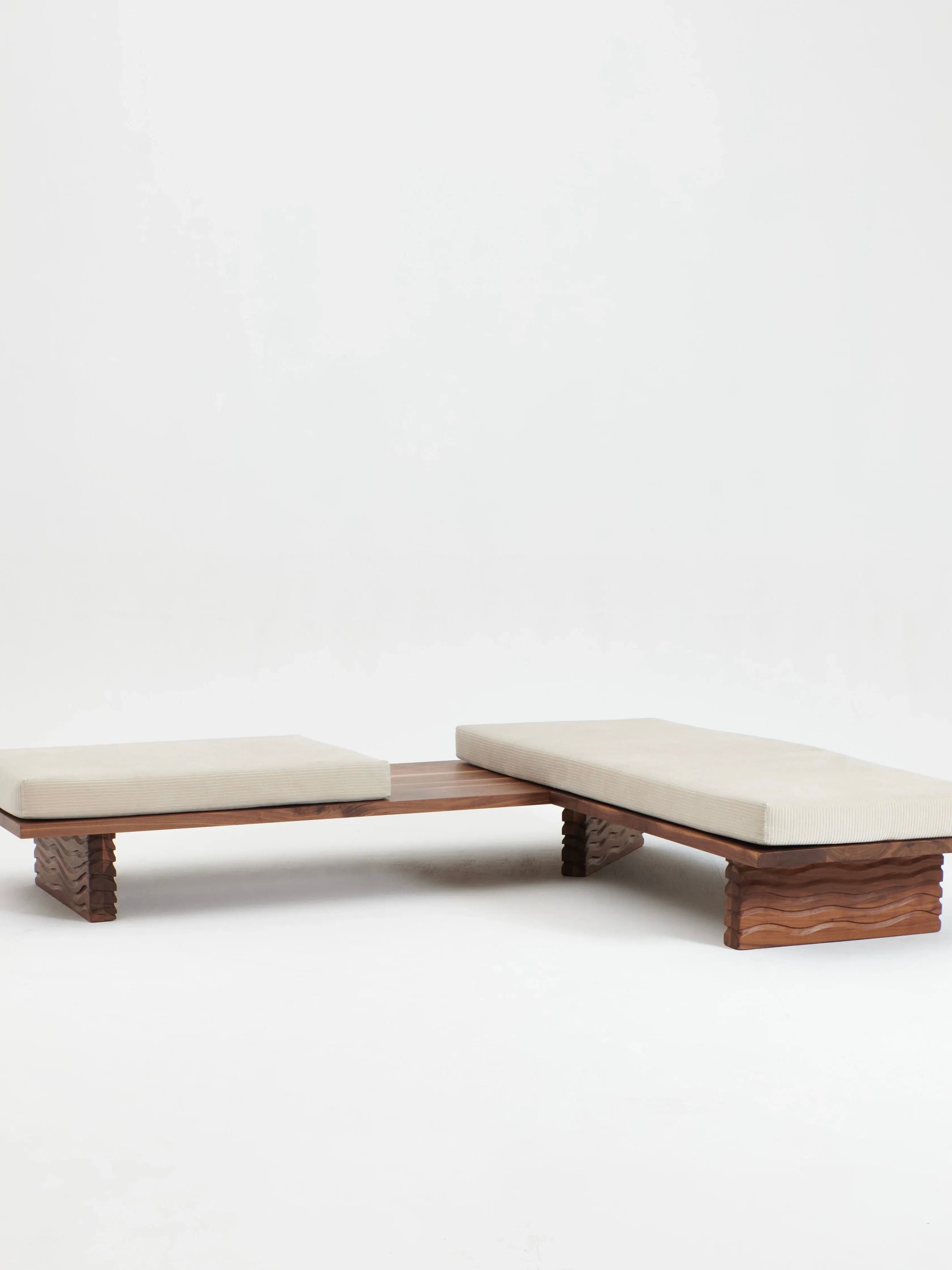 Introducing the Nazaré Daybed L Shape by Project 213A, a contemporary and refined double bench showcasing two beige cushioned seating areas separated by a solid walnut surface in the center. Handmade in Portugal, this piece stands elegantly on intricately carved wooden legs and is presented against a plain white background.