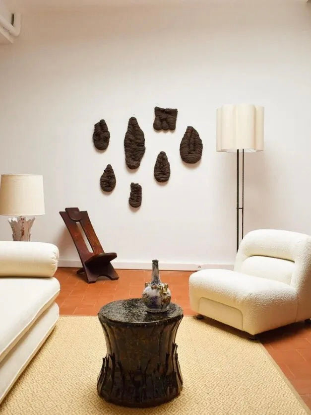 A minimalist living room features white sofas and a textured coffee table on a beige rug. Abstract dark sculptures adorn the white wall alongside the West African Wood Chair, 1960s from Barracuda Interiors. Two floor lamps, one silver and one black, illuminate the space near a wooden magazine rack.