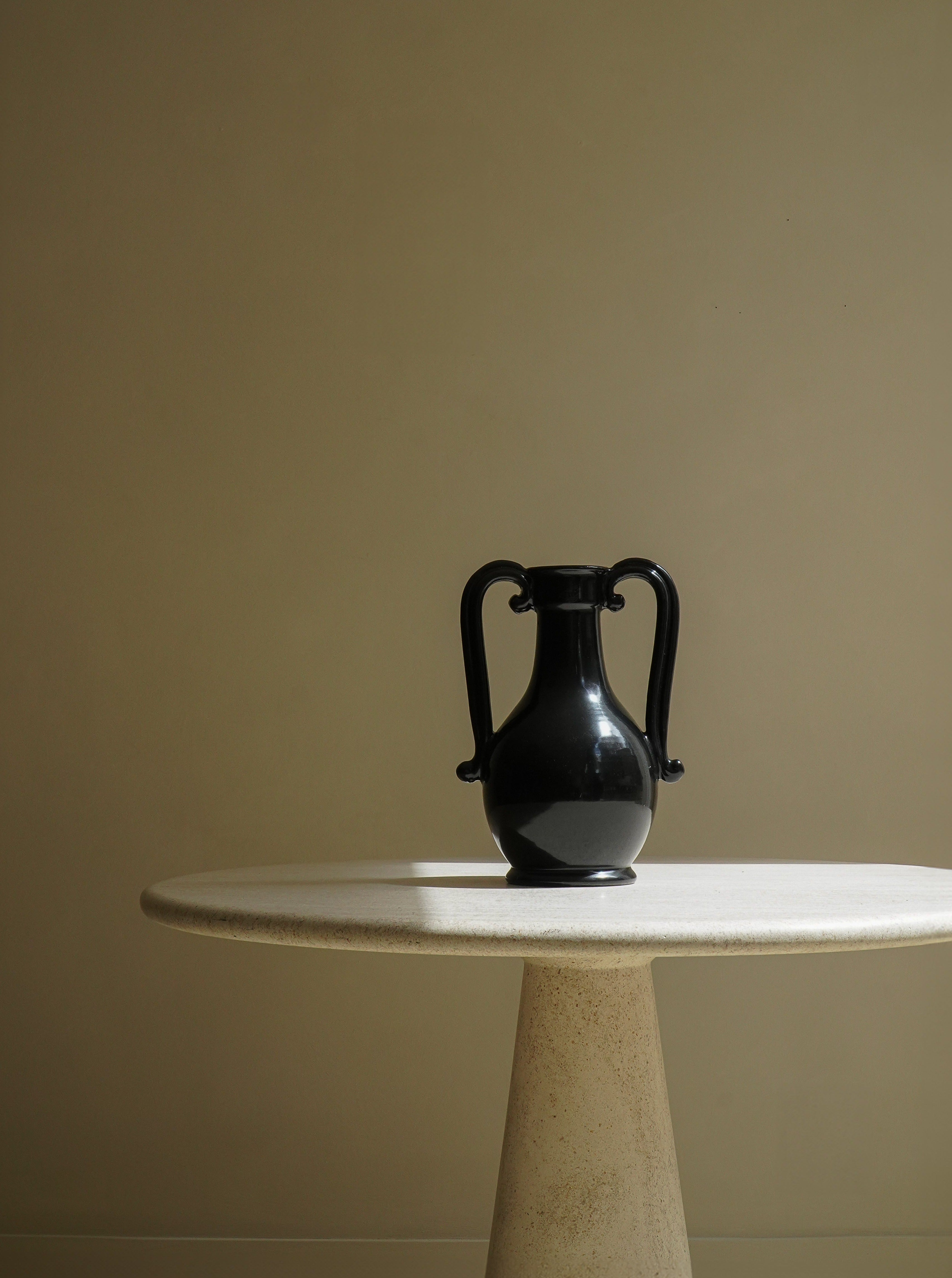 A black two-handled vase, **The Fiasca Vase** by **COSEINCORSO**, showcasing Italian craftsmanship, sits centered atop a minimalist, round, white table with a tapered pedestal base. The scene is set against a plain beige wall, creating a stark contrast and a serene, modern decor composition.