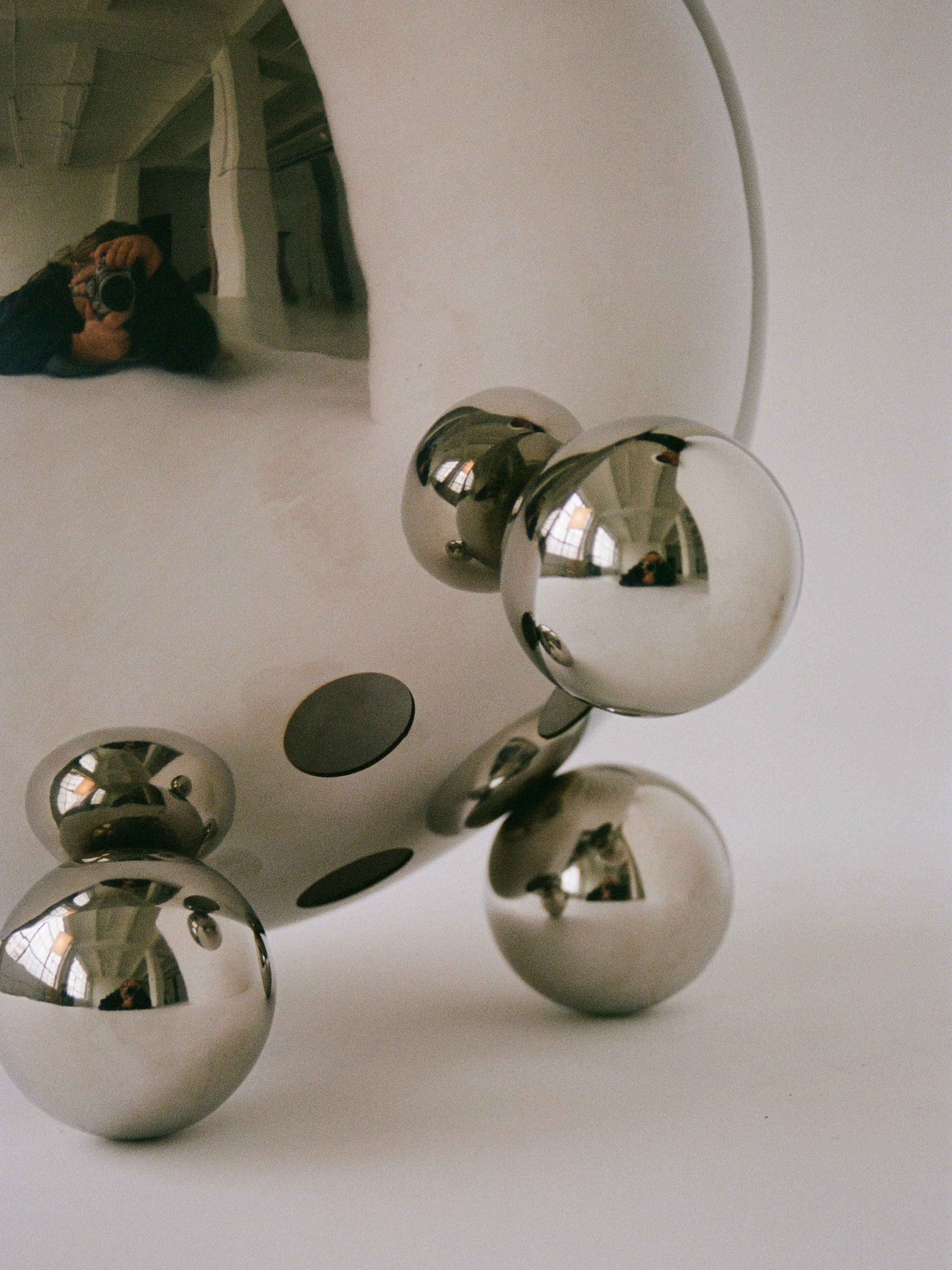 A person captures a photograph in the reflection of the smooth, stainless steel Selene Planter, Large by MUHLY. Composed of various-sized spheres, this sculpture reflects their surroundings and the photographer. The backdrop is minimal and light, featuring faintly visible windows and structural details.