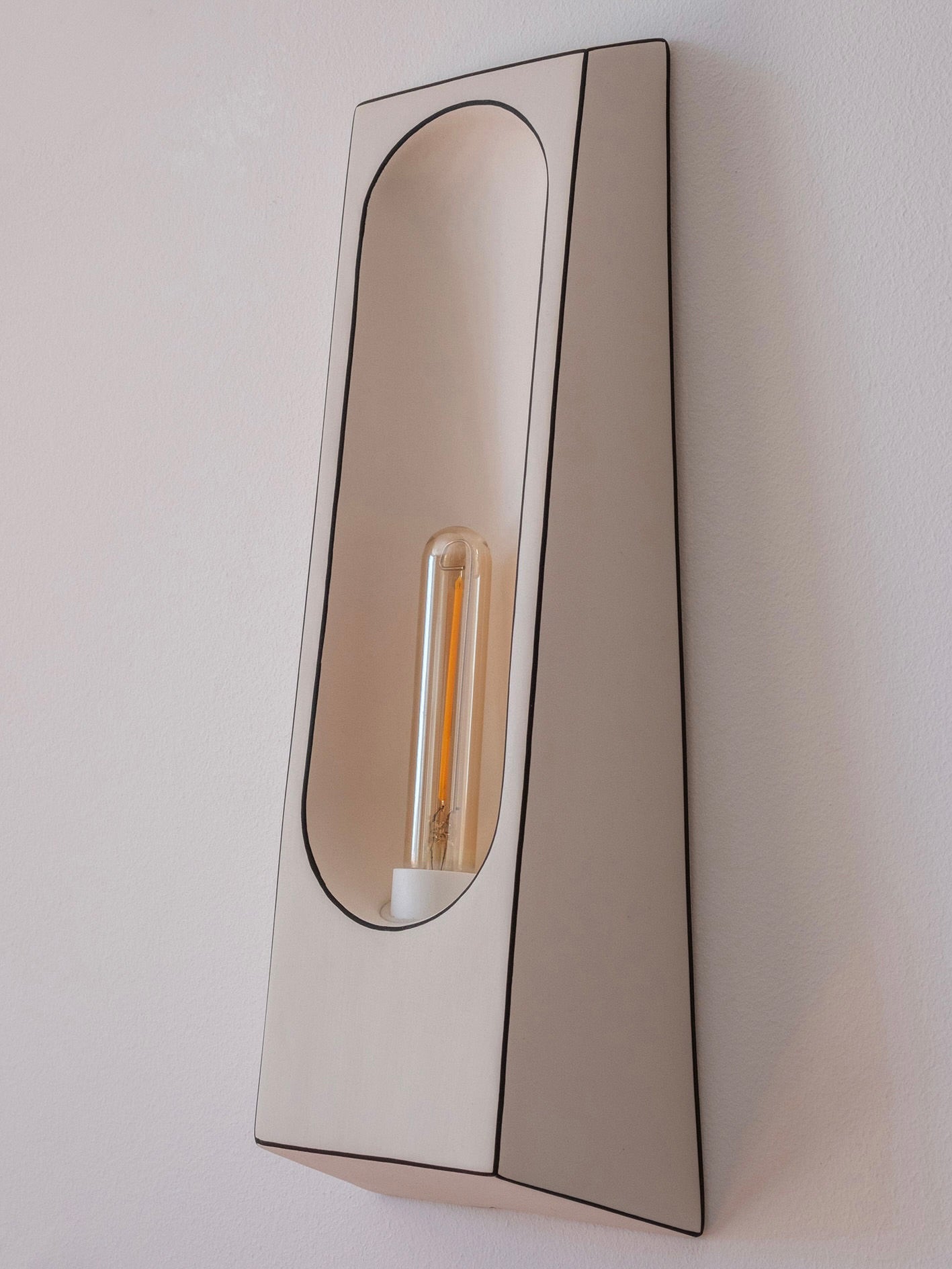 The Alcove Facette Wall Lamp by Violaine d'Harcourt is a modern, wall-mounted accent light featuring a sleek, curved design. Its hand-finished, minimalist beige housing holds a long vertical bulb, casting a soft glow on the wall.