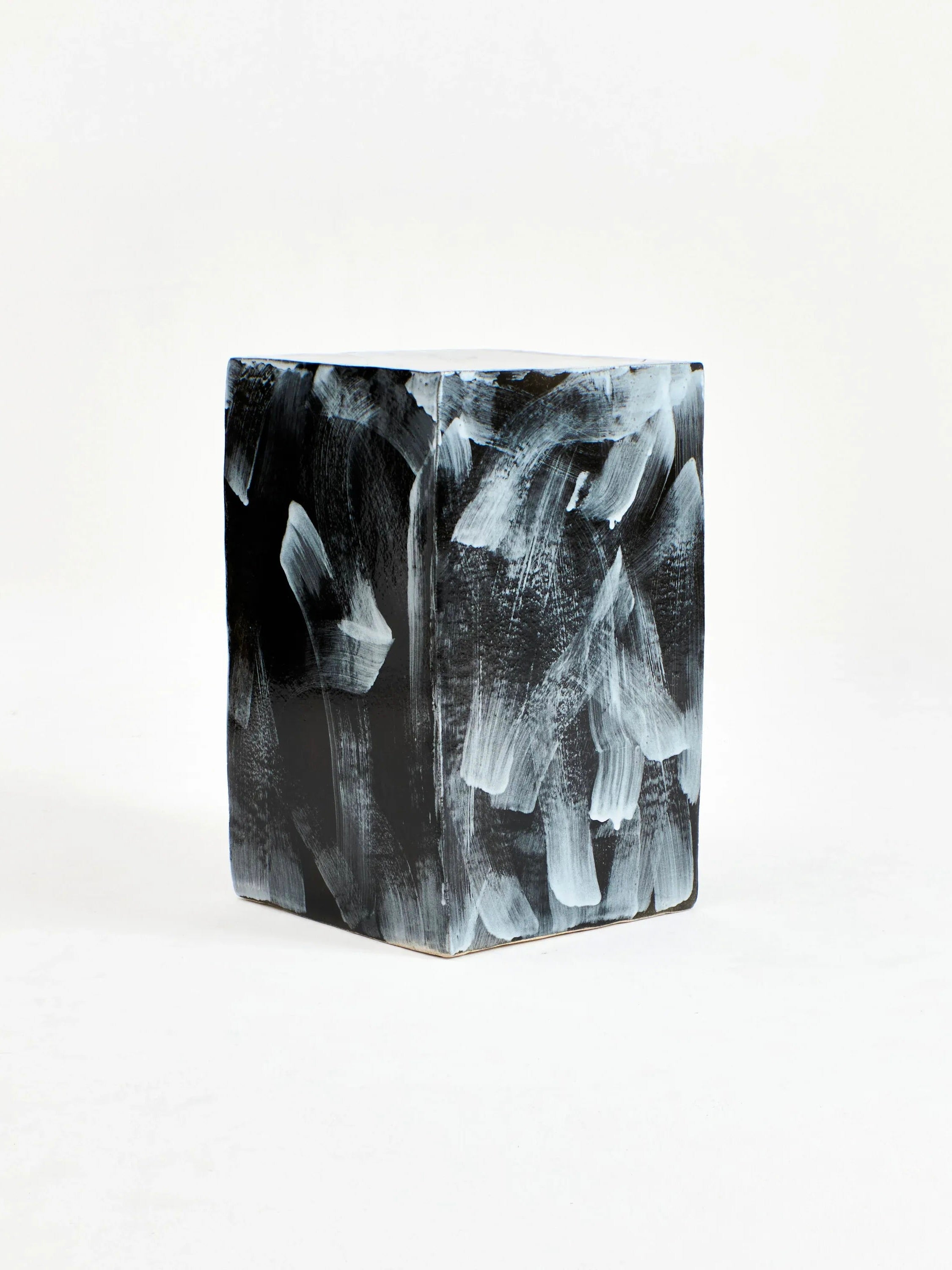 A ceramic side table by Project 213A stands upright against a light background, featuring a black surface adorned with sweeping abstract white brushstrokes. The design’s textured appearance gives the table a modern and artistic look, reminiscent of geometric shapes often seen in handmade pieces from Portugal.