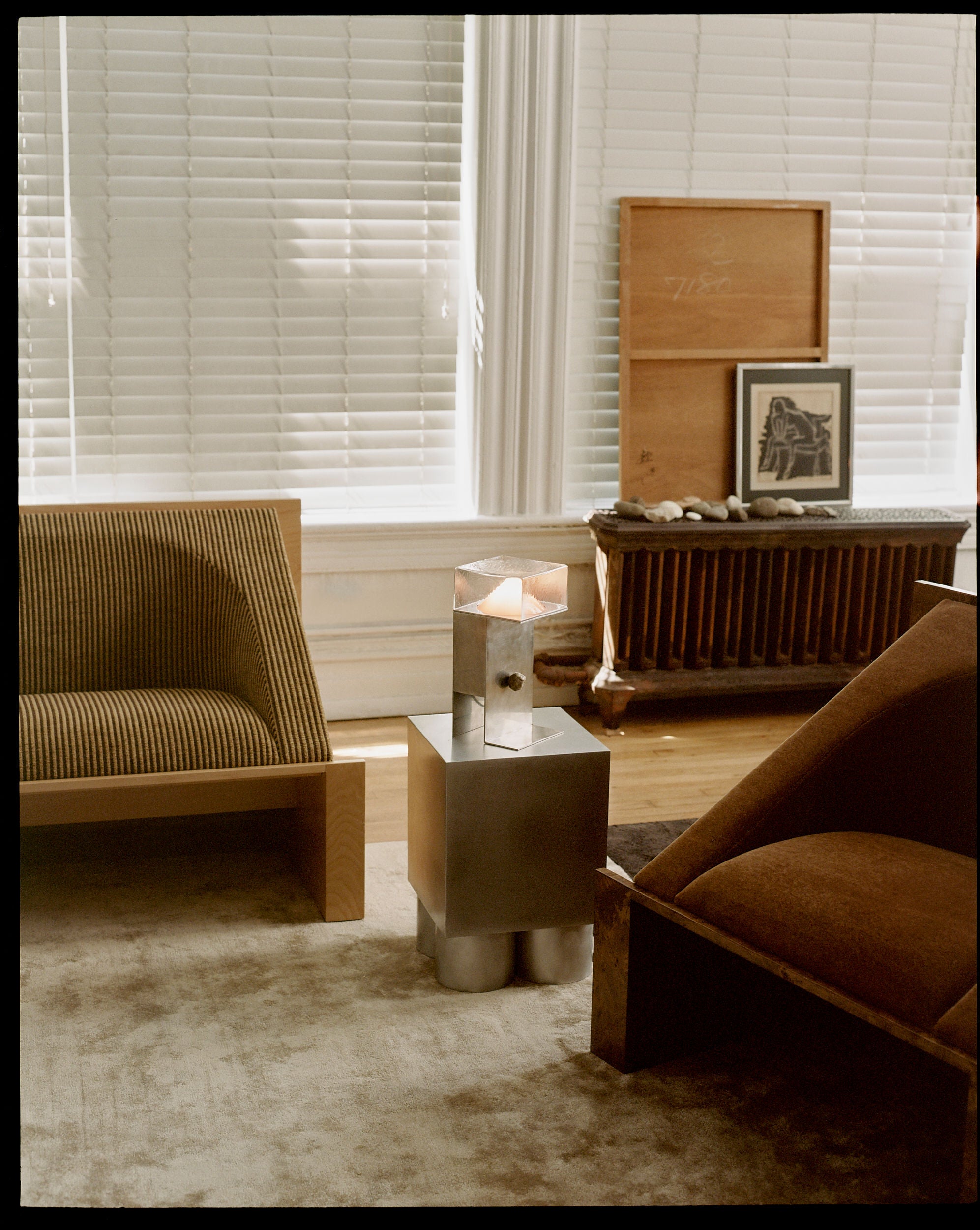 The minimalist living room includes two armchairs—one in brown and a 'Perceptions' Armchair by Known Work with stripes. A metal side table with a lit candle adds warmth. Large white blinds and an art-accented radiator enhance the inviting space, complemented by a soft, neutral-toned rug.