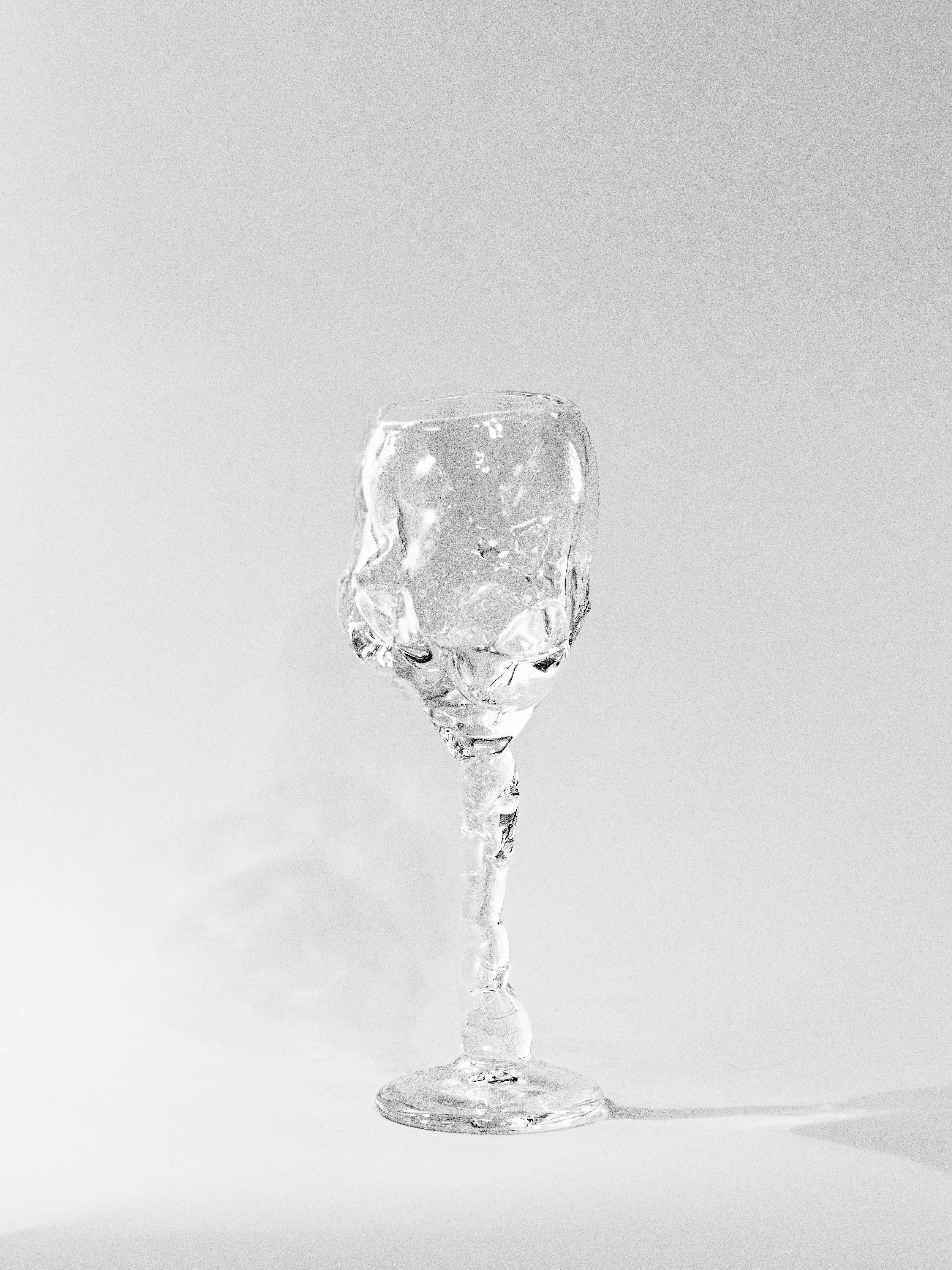 The Hand Blown Freeform Wine Glass by Szkło Studio rests on a white surface with a light gray background. Made from transparent sodium glass, its abstract, twisted form offers an artistic and unique aesthetic akin to Polish craftsmanship.