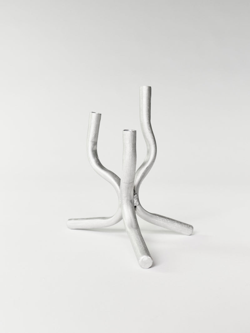 The Mini Candelabra by Six Dots Design is a minimalist metallic sculpture handcrafted in a North London workshop, featuring four interwoven curved tubes extending upwards like abstract tree branches, set on a plain white background.