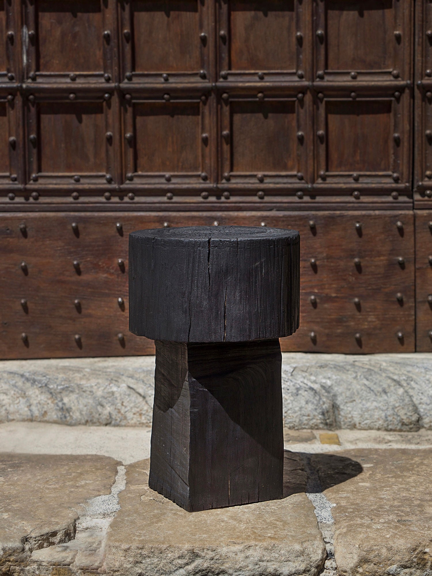 The Touch With Eyes Burnt Black Coffee Table/Stool, expertly handmade with a textured, natural finish, stands on stone before an ornate wooden door with metal studs. Its cylindrical top and square base beautifully complement the rustic surroundings.