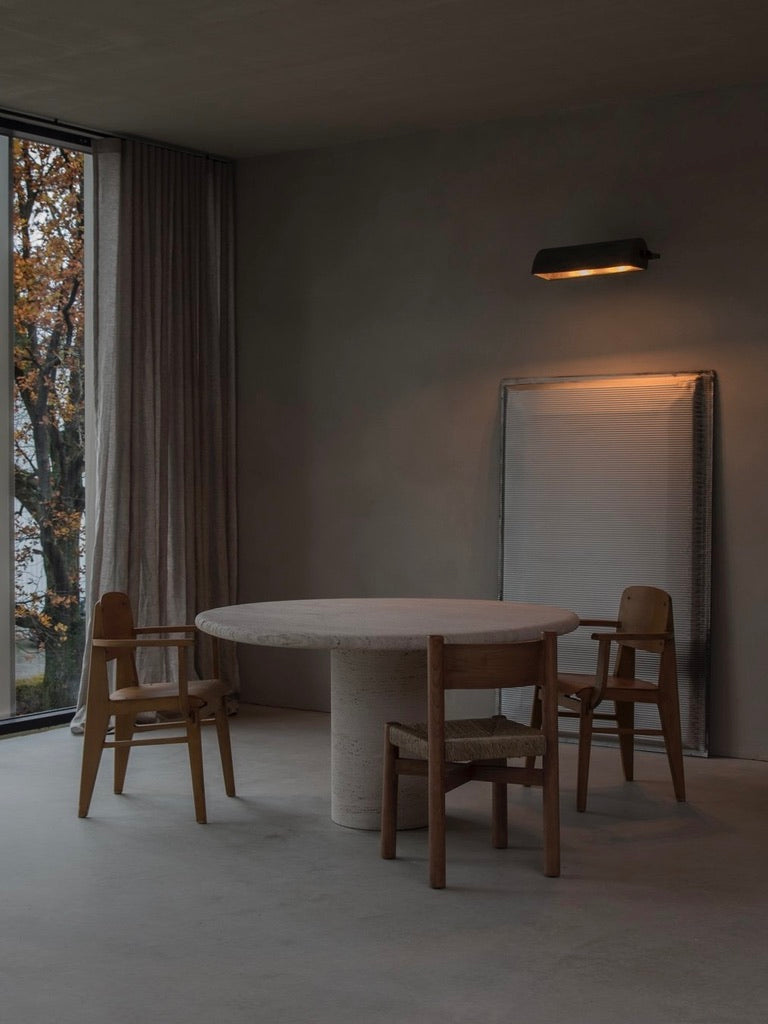 A minimalist room features a round stone table and three 'Les Allues' Straw Chairs by Bicci de' Medici. A large window with drawn curtains reveals autumn foliage. An abstract painting leans against the wall under a modern lamp, evoking a cozy atmosphere.