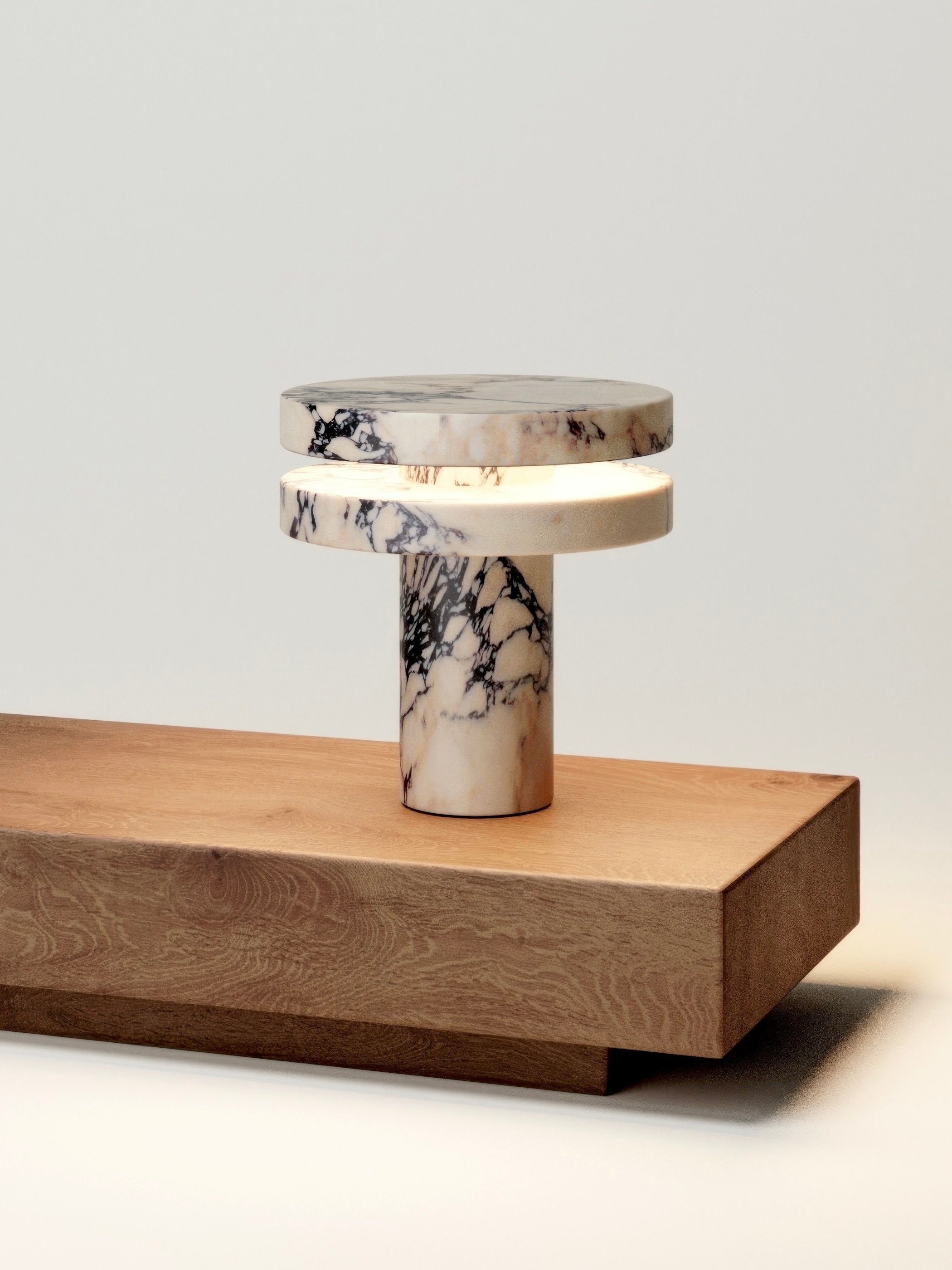 The 'M_014' Marble Table Lamp by Monolith features a cylindrical base and layered design with a black and white marbled pattern. It sits on a wooden surface, echoing brutalist style while emitting a soft LED glow between the layers.