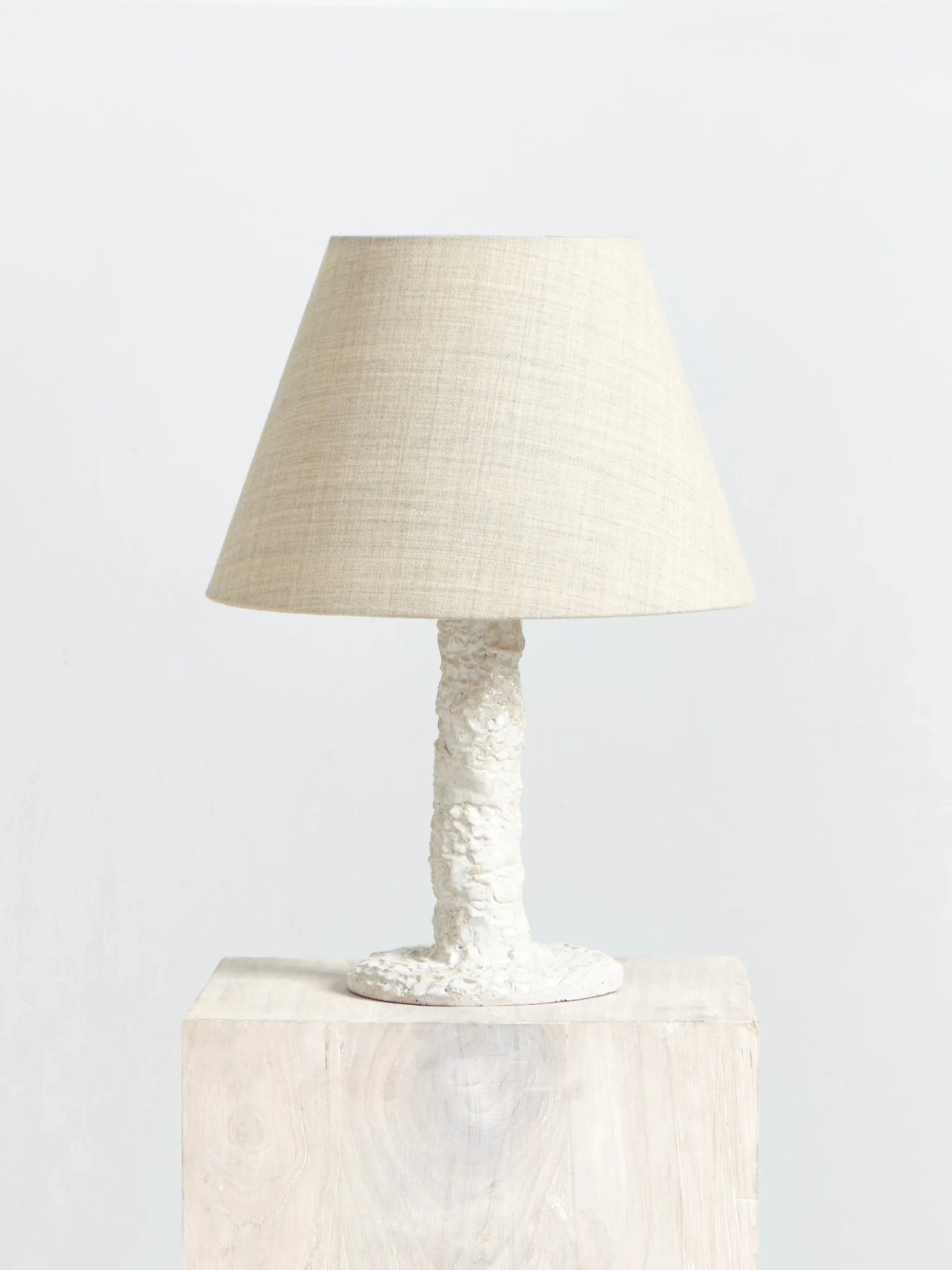 The Casa Bedside Light by Project 213A, featuring a handmade table lamp with a textured white base and a beige fabric conical shade, is displayed on a light wooden surface against a plain white background.