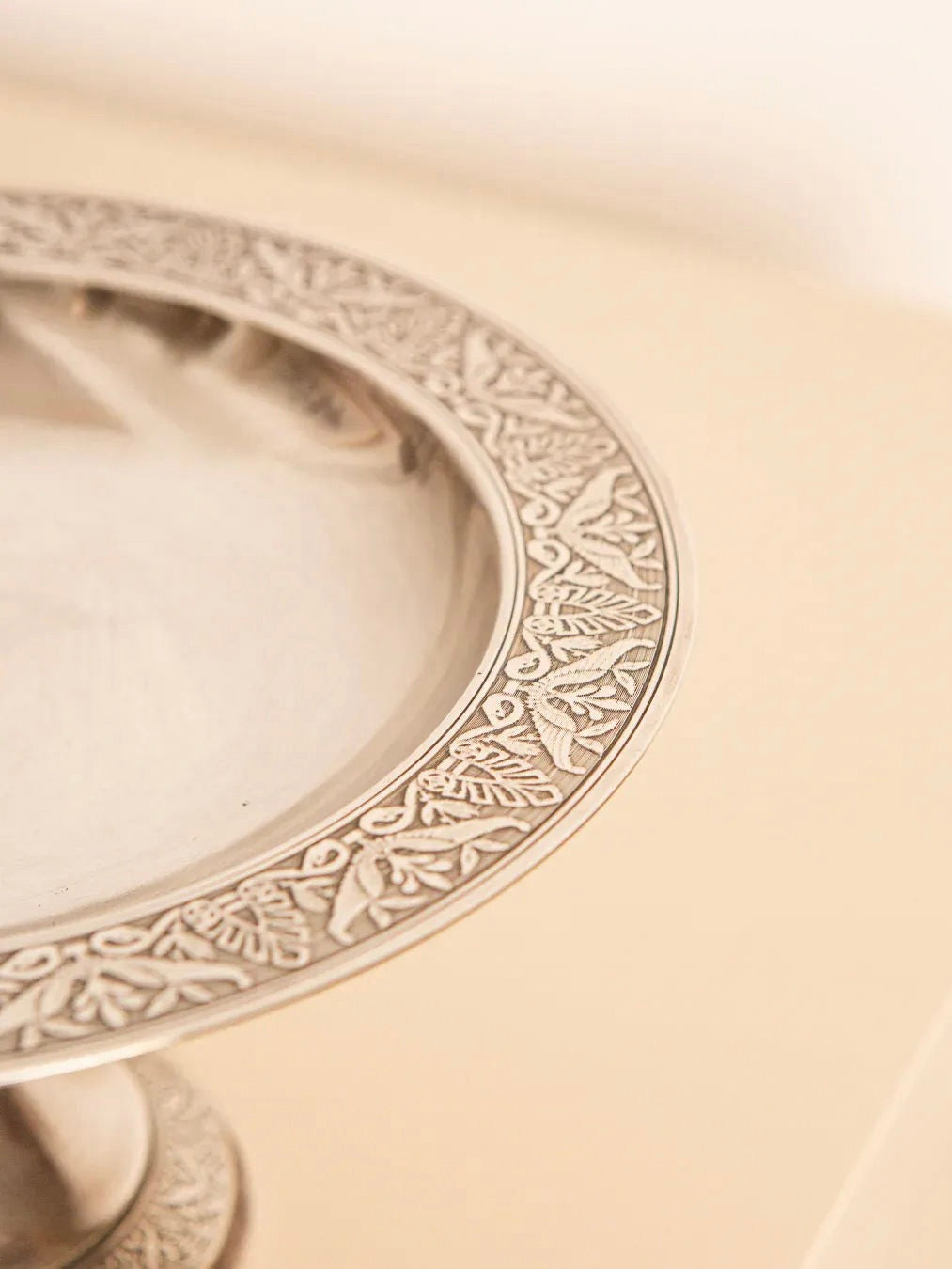 A close-up features the Treaptyque Engraved Silver Plated Fruit Basket 60's, showcasing its ornate design with intricate patterns along the rim against a neutral background. Its reflective surface hints at a retro style reminiscent of 1960s design, possibly serving as both a decorative tray and pedestal.