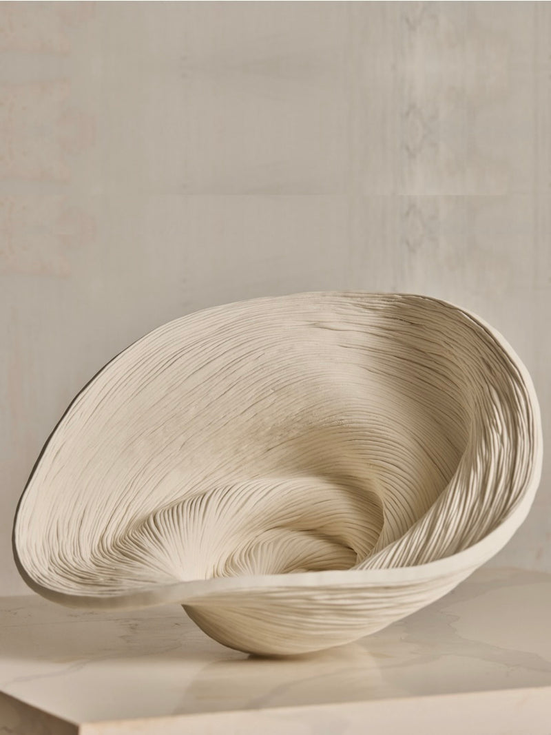The Soucy Shop's Porcelain Bowl, an elegant sculptural piece crafted from hand-rolled porcelain, features a swirling design and textured creamy surface akin to a seashell. Against a muted wall, its unique curves make it truly one-of-a-kind.