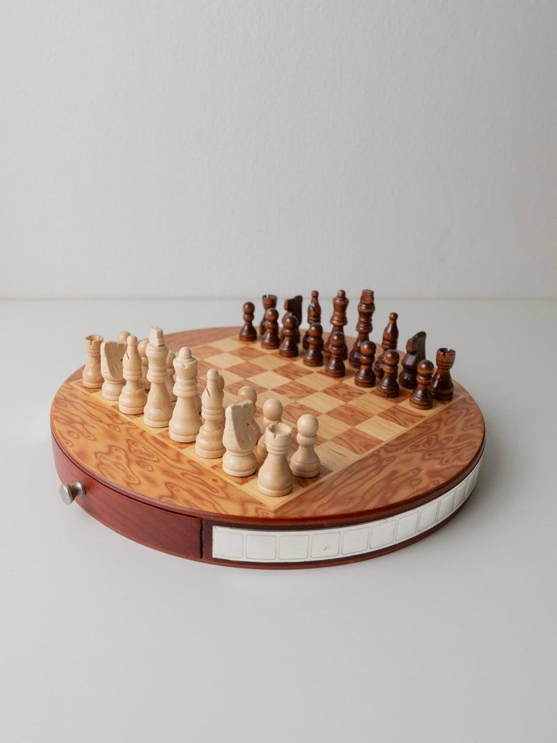 Bottega Jacobs' Mid Century Design Chessboard from 1960s Italy features a round vintage board with a drawer and hand-carved wooden pieces. Its natural wood grain finish enhances its charm, as it rests elegantly on a white surface against a plain wall.