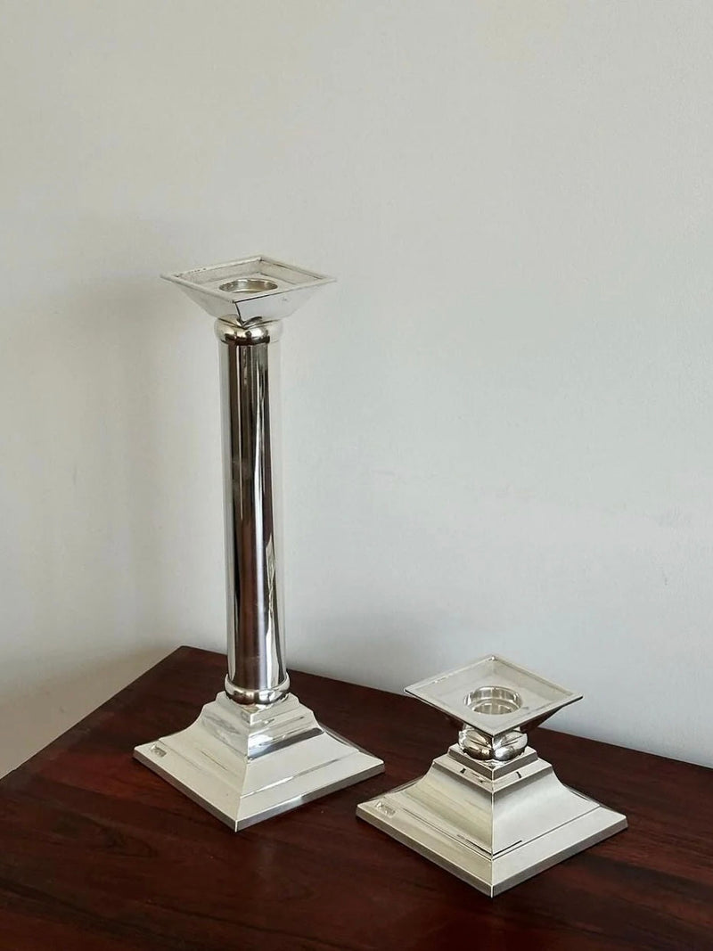 A photo shows two vintage German Candle Holders Pair by Aesthete Label on a wooden surface. The taller holder features a slender, classical column style, while the shorter one is more compact. Both candle holders have square bases and top holders designed for holding candles.