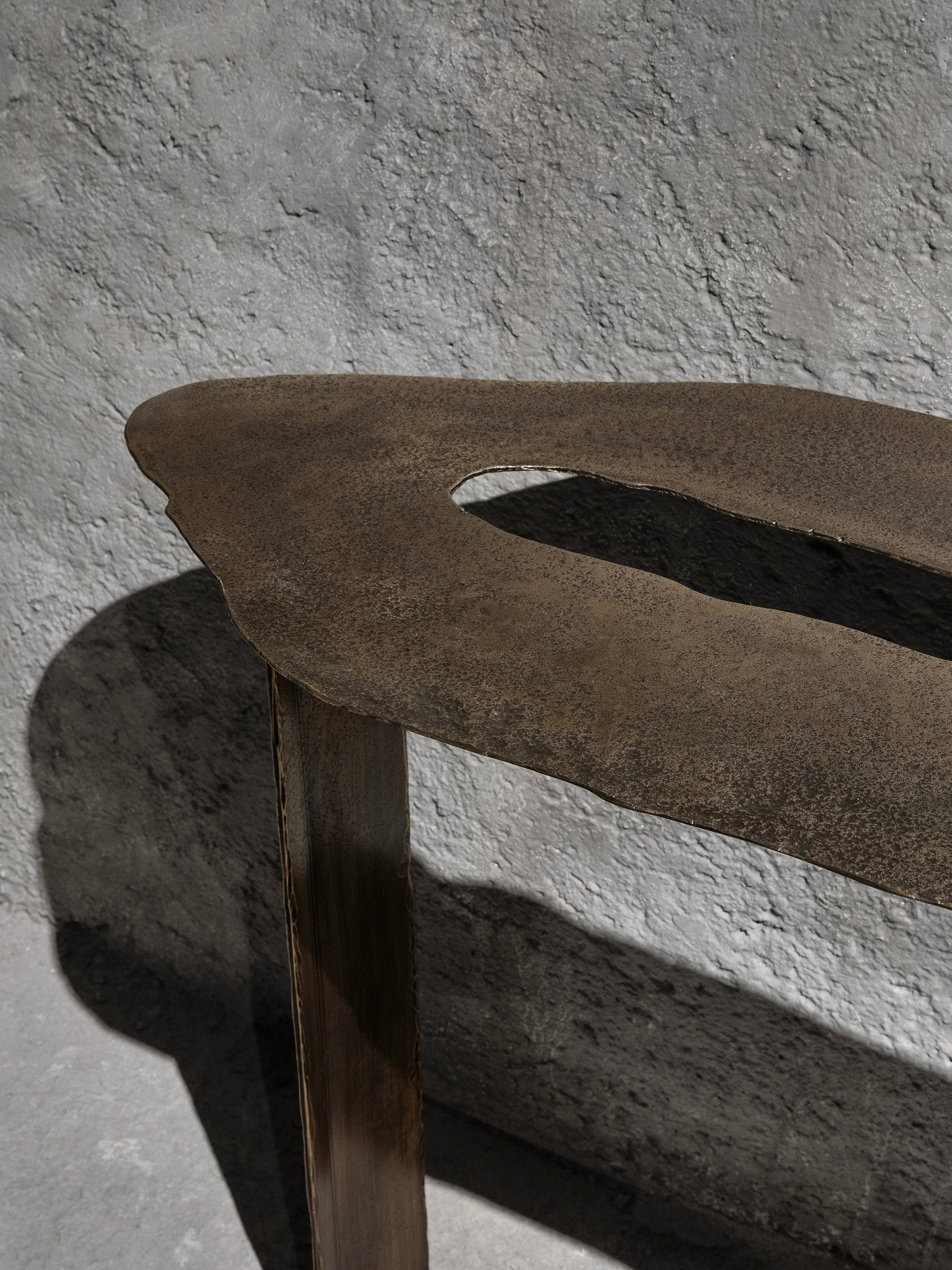 Close-up of the Muisca Console Table by Ombia Studio, featuring an abstract, textured metal design with a brass finish and a rough oval cutout. It stands against a concrete background, casting a shadow on the wall for added depth and contrast.