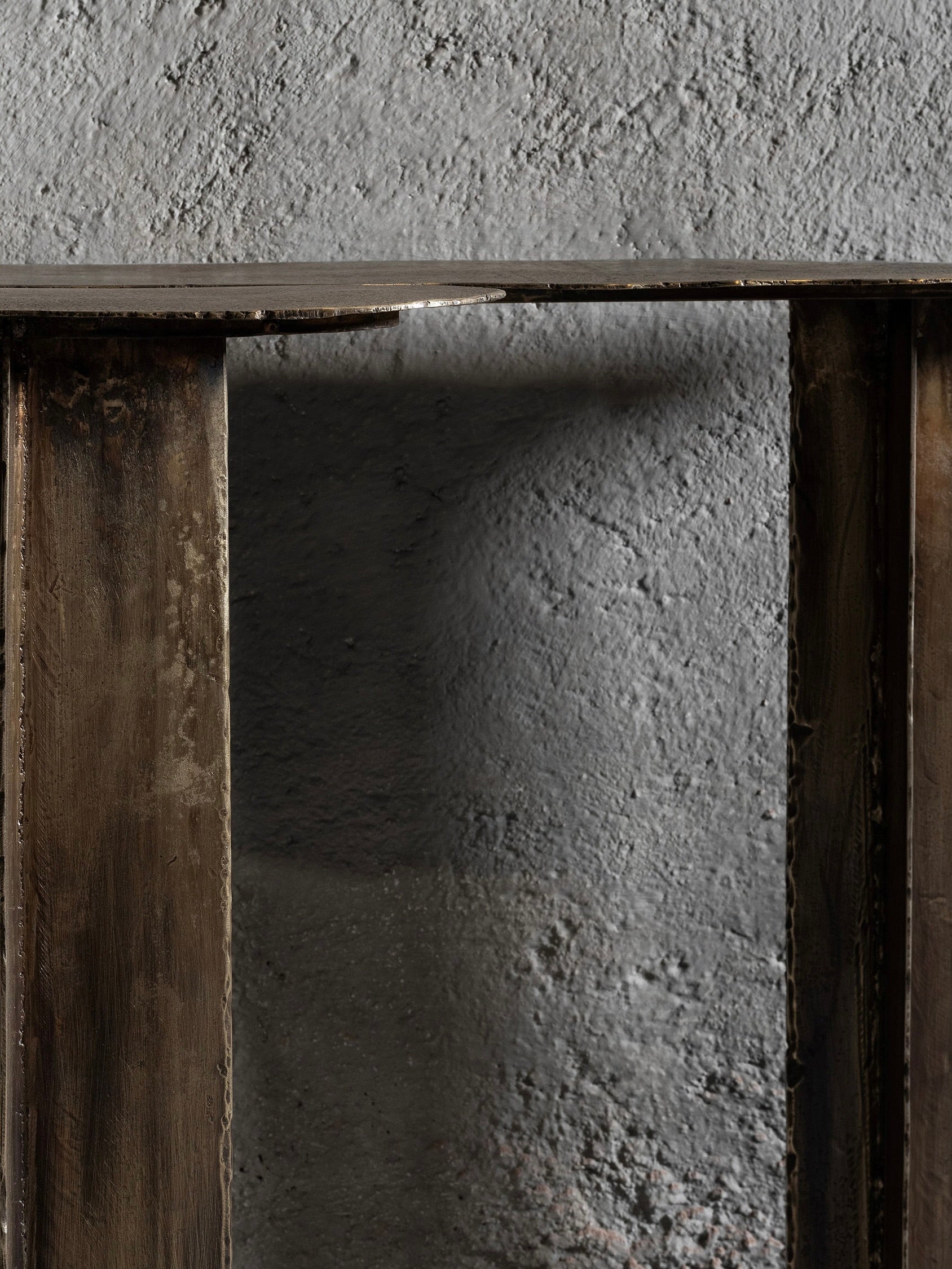 Close-up of the Ombia Studio Muisca Console Table, featuring a brass textured finish against a rough gray concrete wall. Its metallic surface and vertical supports show wear, adding a vintage, Muisca-inspired industrial feel to the minimalist scene.