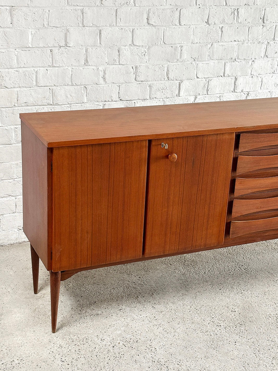 The Introverso Italian Modernist Teak Sideboard, inspired by Vittorio Dassi and the 1960s aesthetic, has angled legs, a left-side cabinet with a round handle, and four horizontal drawers. It’s set against a white brick wall on a concrete floor.