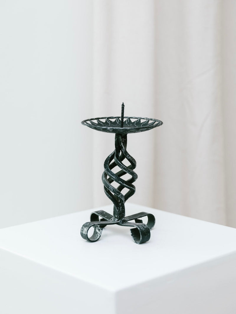 The Forged Candleholder – Brutalist Design 1950s by Bottega Jacobs, featuring an intricate spiral design, is showcased on a white pedestal with a light-colored curtain backdrop, embodying a subtle Brutalist charm.