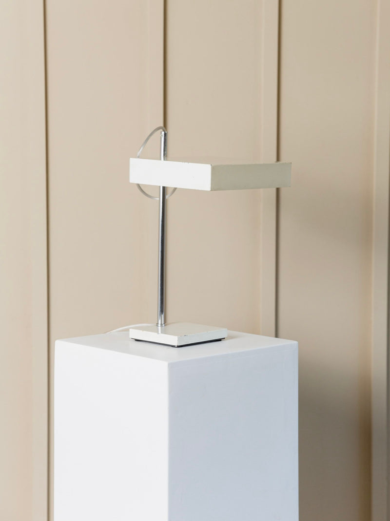 A contemporary white sculpture with a mid-century modern vibe is showcased on a tall pedestal against a beige, paneled wall. The piece boasts geometric shapes, featuring a vertical rod and horizontal element that mirror the sleek elegance of the Kaiser Leuchten Table Lamp by Klaus Hempel from Bottega Jacobs.