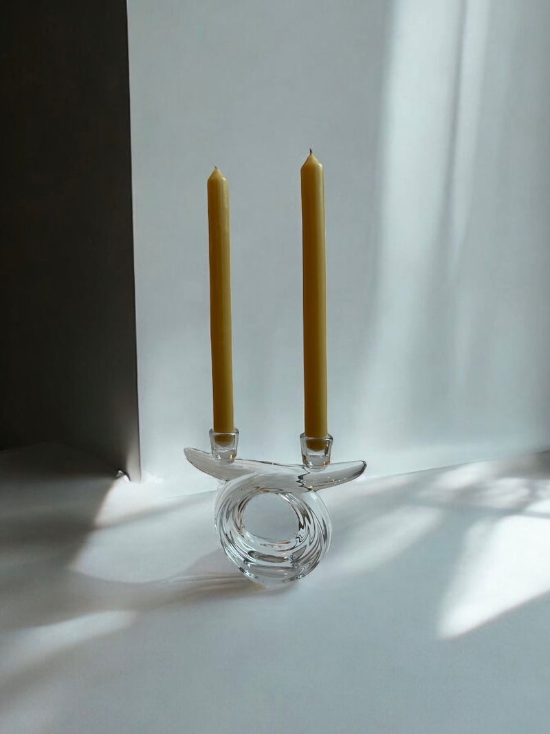 The Crystal Candle Holder 1960s by Flock Studio, with its modern sculptural design, holds two unlit yellow taper candles against a softly lit minimalist backdrop, exuding mid-century elegance reminiscent of 1960s French crystal.