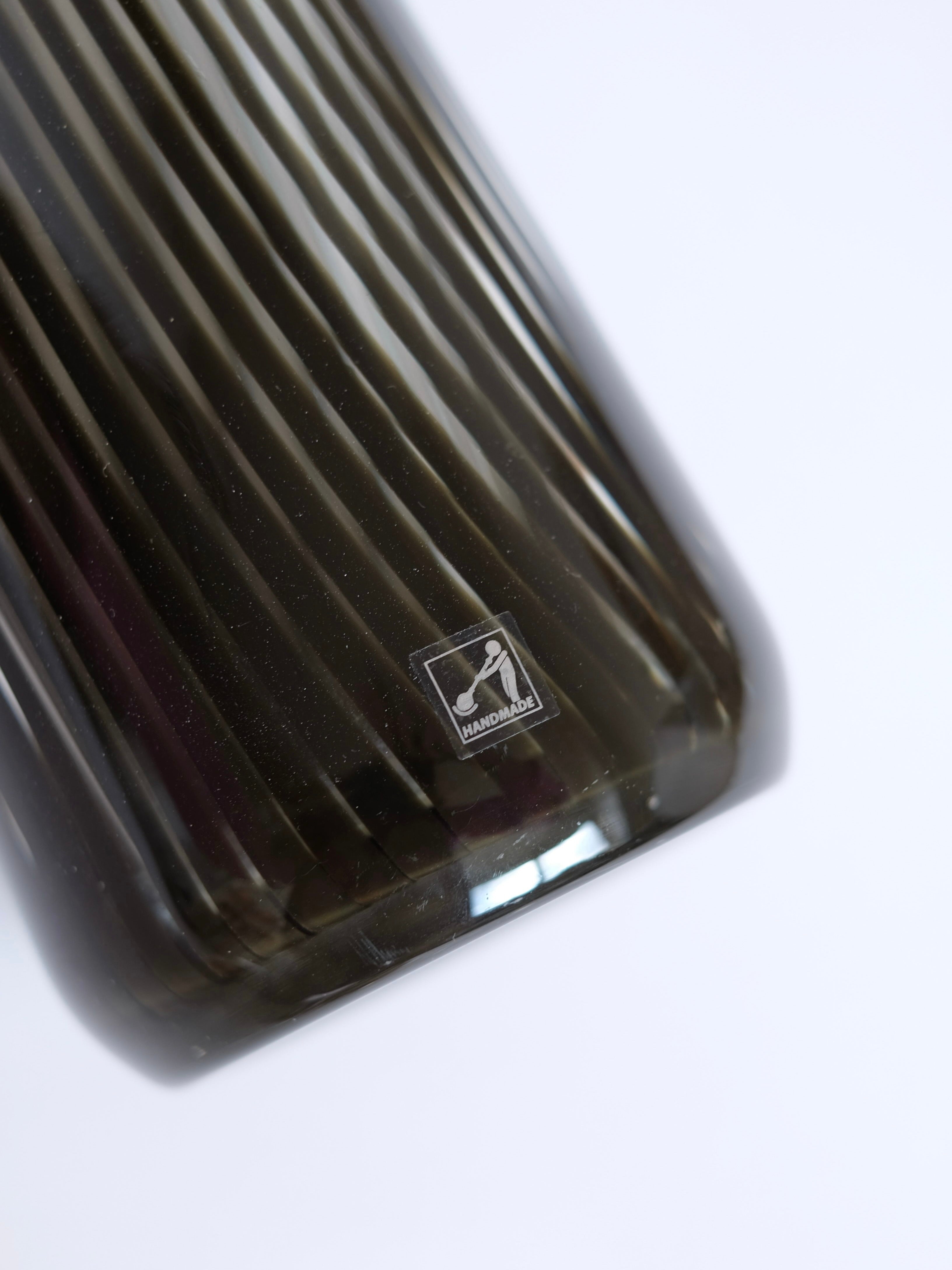 Close-up of the Italian Murano Vase, 1970 by septembre studios, a dark, ribbed glass piece with a small square label reading "HANDMADE" on its side. The smooth, shiny surface reflects the delicate elegance of vintage Murano glass against the plain white background.