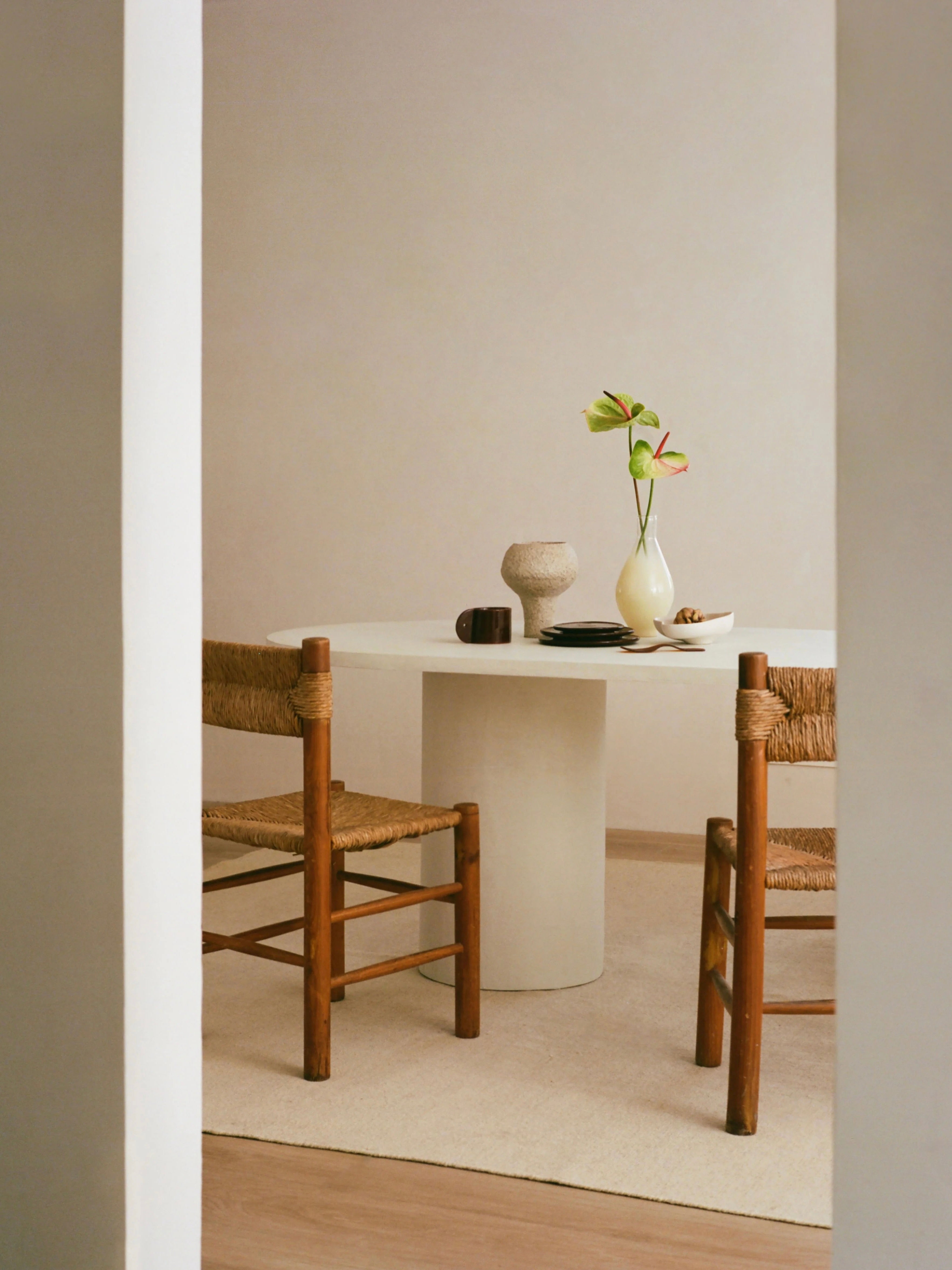 In a minimalist dining area, the contemporary Reye Dining Table by MarlotBaus is paired with two handcrafted woven wooden chairs and a flower-filled vase. Neutral walls complement the serene and simple vibe.