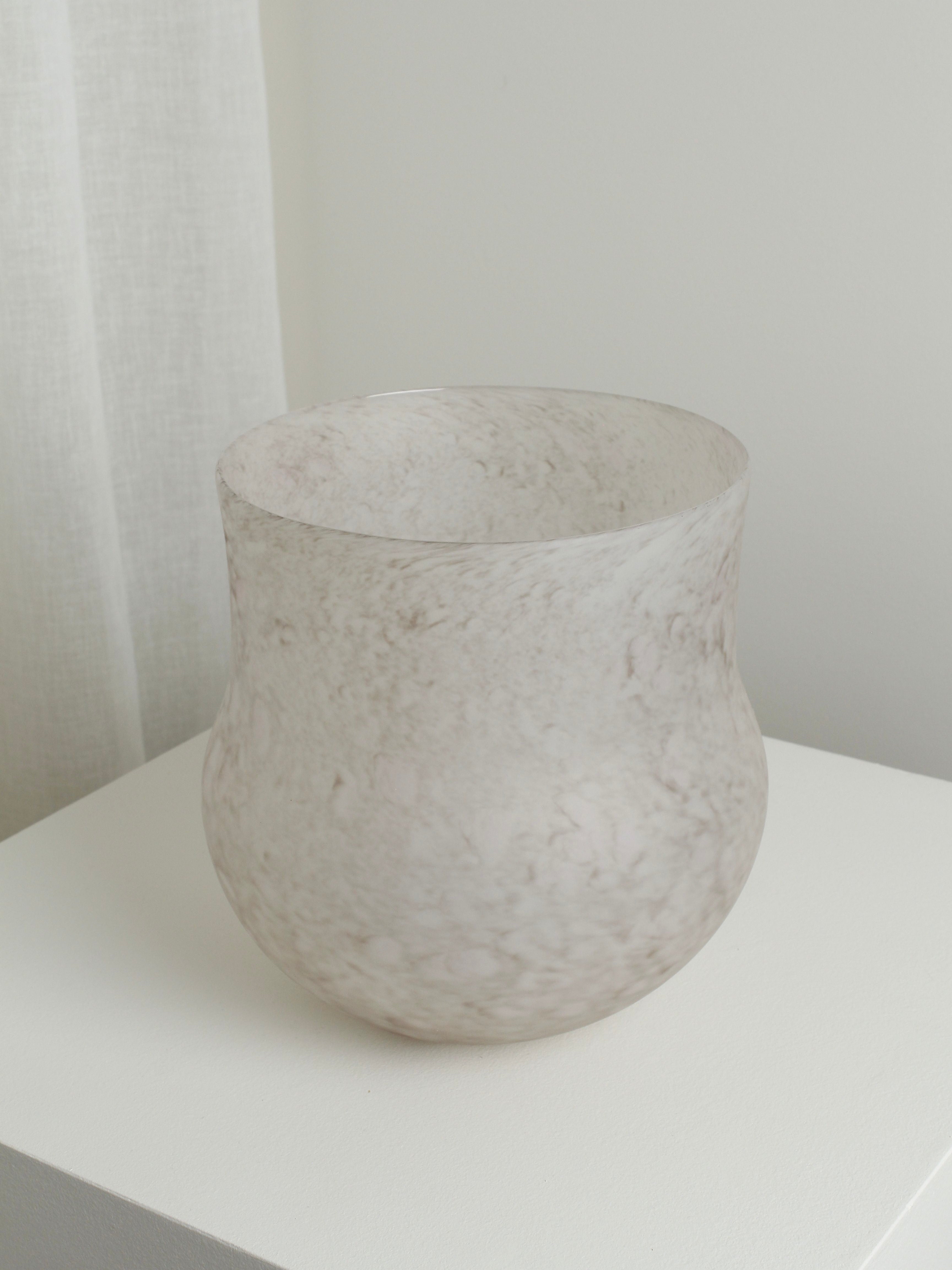 A matte, translucent vase with a marble-like pattern sits on a white pedestal. The Verre d'Onge XLarge Vase in Feathered Taupe, featuring a rounded lower portion and slight flaring at the top, is mouth blown. It is placed in a minimalist room with light-colored walls and curtains matching its Feathered Taupe design in the background.