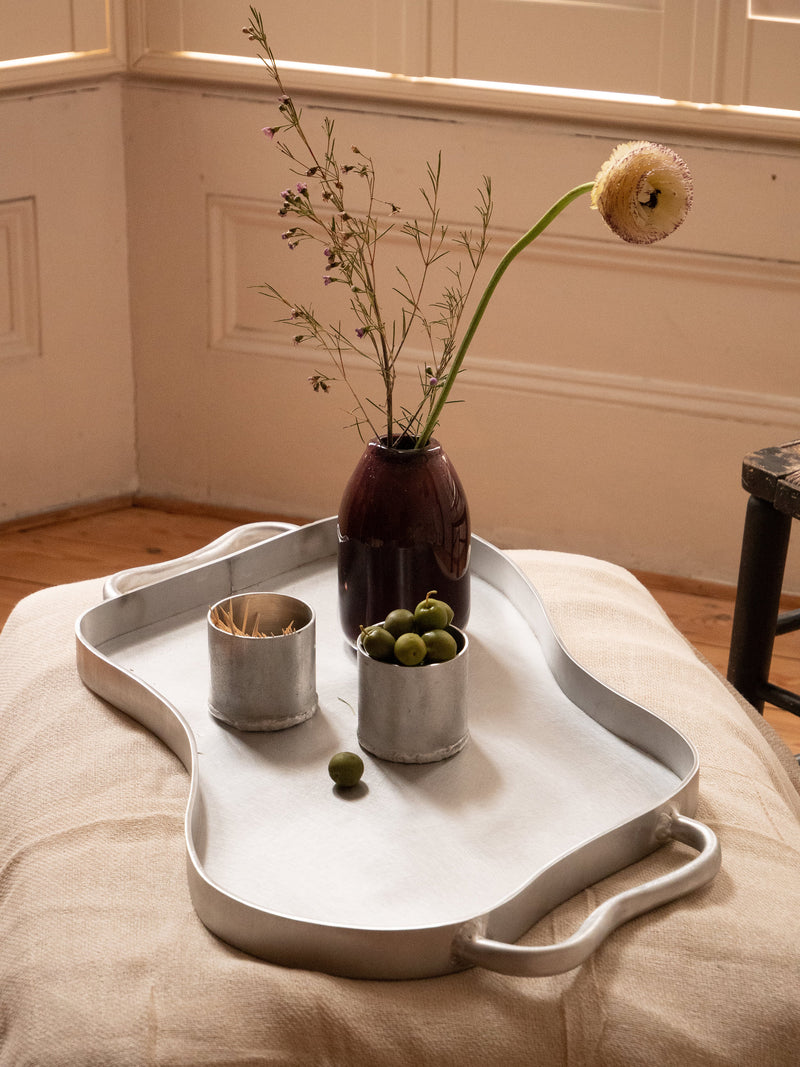 The Six Dots Design Serving Tray holds a dark vase with wildflowers, a tin cup of toothpicks, and another with green olives. The tray rests on a beige cushion by a sunlit window in North London.