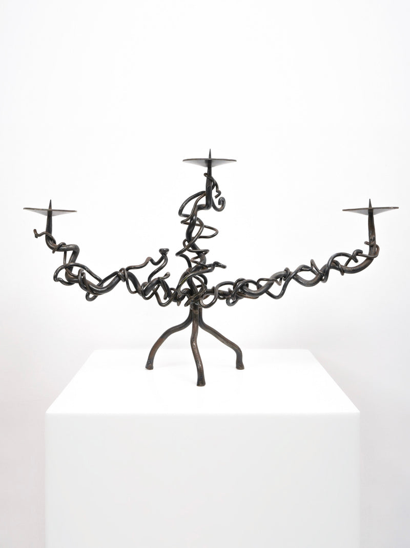 The Tom Bogle Brutalist Iron Candelabra features twisted black iron with intricate, vine-like arms and three candle holders, hand forged and shown on a white pedestal against a plain white background.