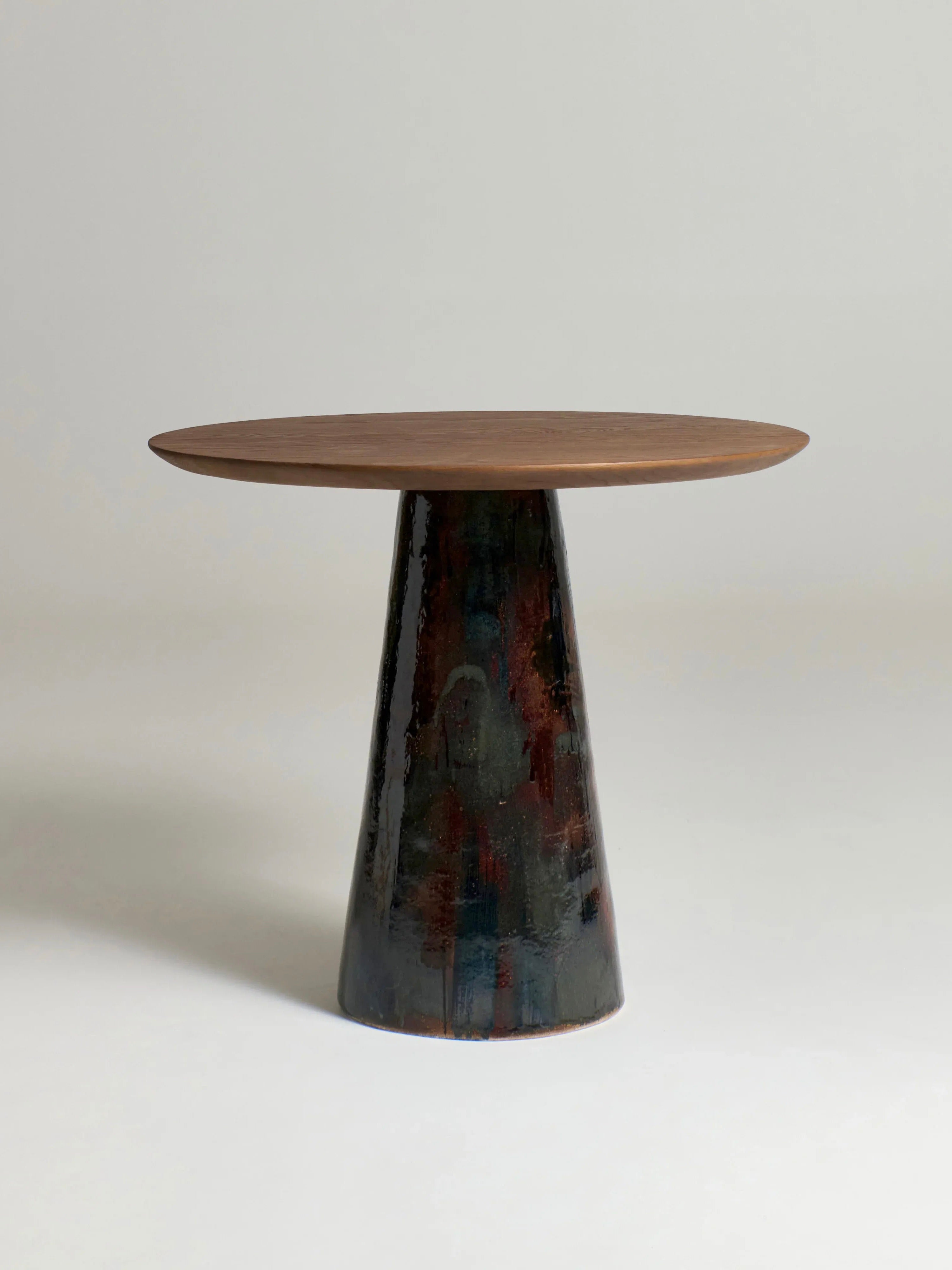 The Porto Belo Table by Project 213A features a round wooden surface with a rustic finish, elegantly supported by a wide, tapered, hand-sculpted ceramic base that adds texture and intrigue against its plain, light-colored backdrop.