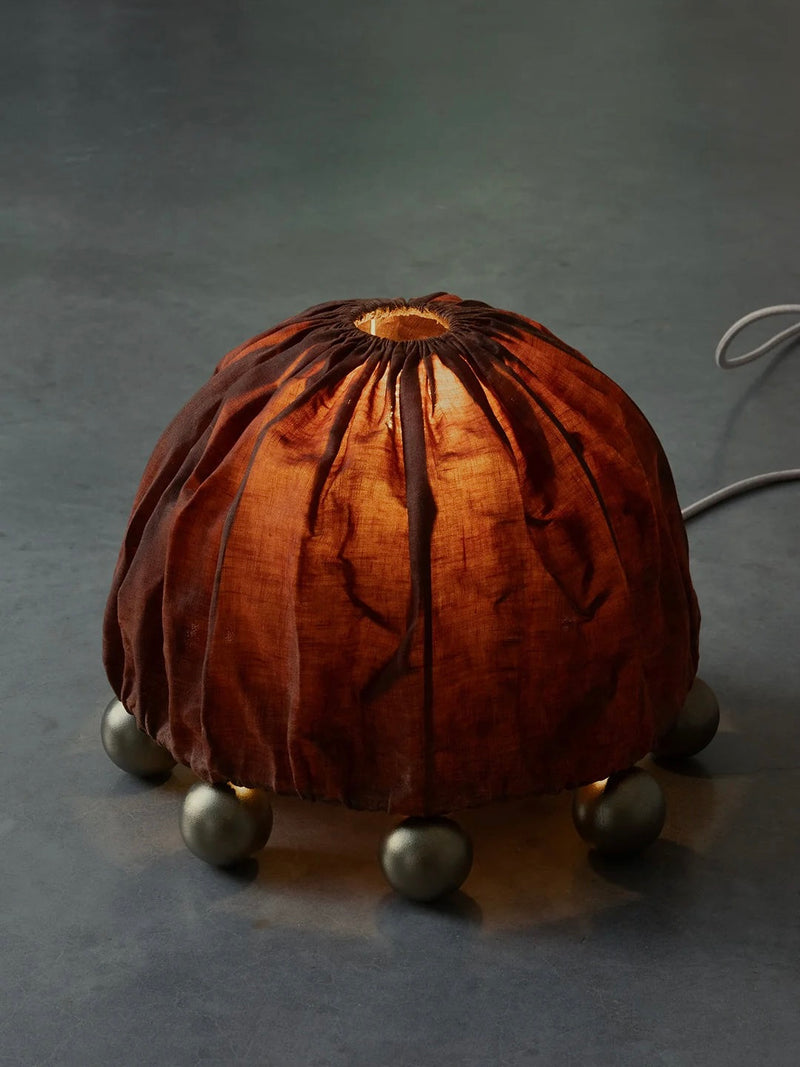 The Kaytar ALTER Table Lamp features a wrinkled, beeswax-coated linen shade resembling a pumpkin. Resting on six small brass legs on a dark surface, this handcrafted lamp emits a warm, soft glow.