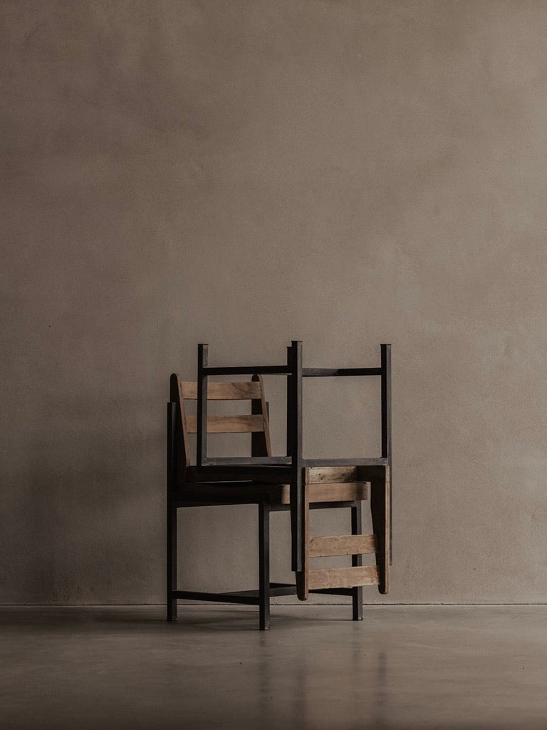 Two Bicci de' Medici Pierre Jeanneret - Authentic PJ-SI-65-A chairs are stacked against a beige wall. They feature dark frames with teak wood slats for seats and backrests, forming a minimalist, shadowed composition on the gray floor.