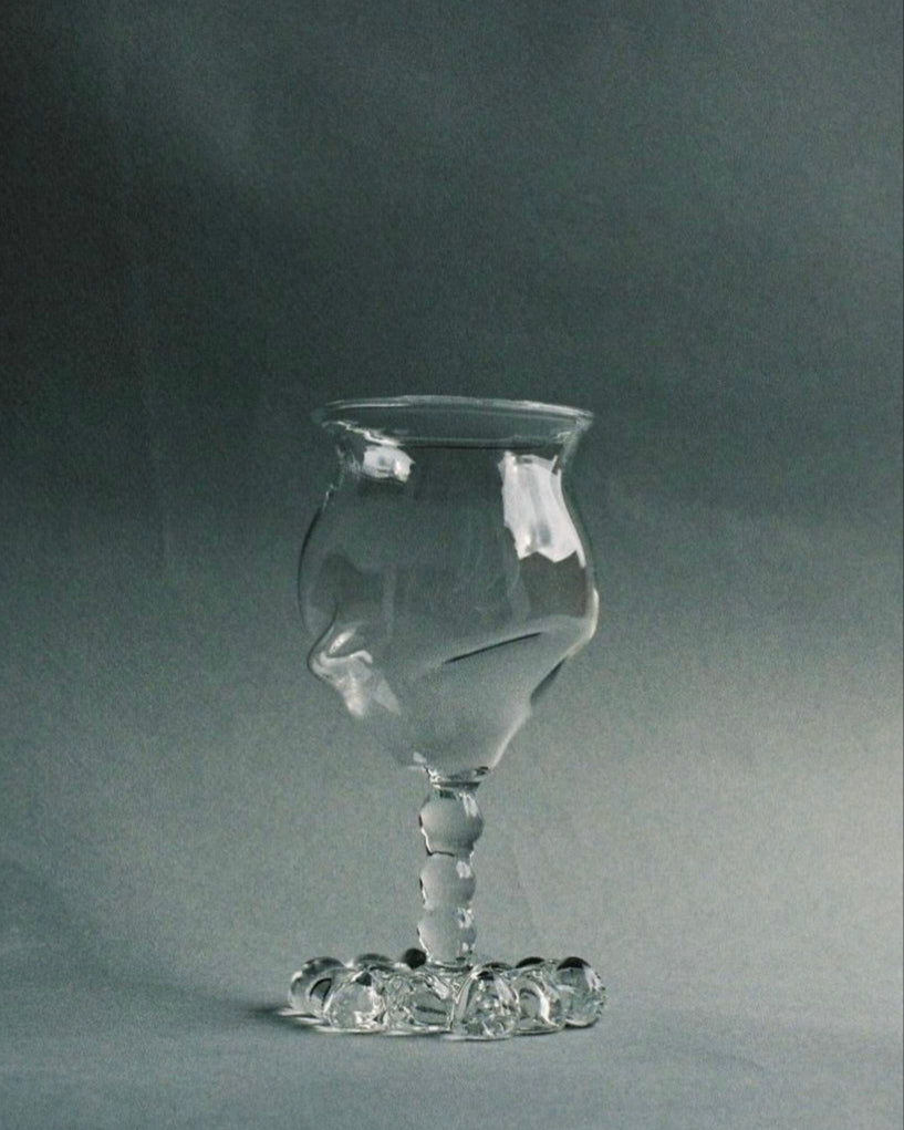 A clear, intricately designed hand-blown wine glass goblet from the "Mouthblown Wine Glass Set" by Justine Menard sits against a plain gray background. This sophisticated wine-drinking goblet features a unique twisted stem and an artistic, slightly asymmetrical bowl. The durable glass base of the goblet is adorned with small circular accents.