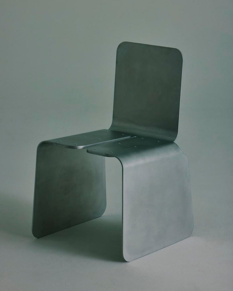 The LESORR Rounded Edges Chair is a minimalist brushed aluminum chair with a matte finish, broad flat surfaces, clean geometric lines, and gently rounded edges. Its seat and backrest are crafted from single bent sheets.
