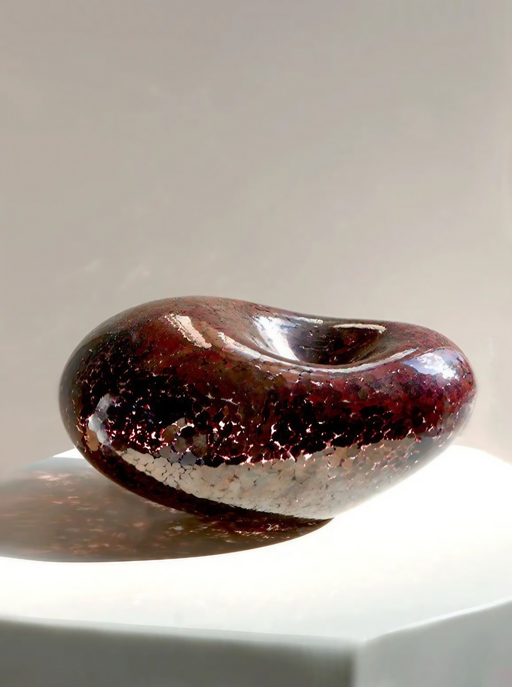 A hollow, dark burgundy glass sculpture named "Lucky Bean in Copper" by Vanderohe Curio sits on a white pedestal. The piece showcases exquisite craftsmanship with its smooth, rounded form and shiny, textured surface, boasting an organic, pebble-like shape and contemporary design that reflects light to cast subtle patterns around it.