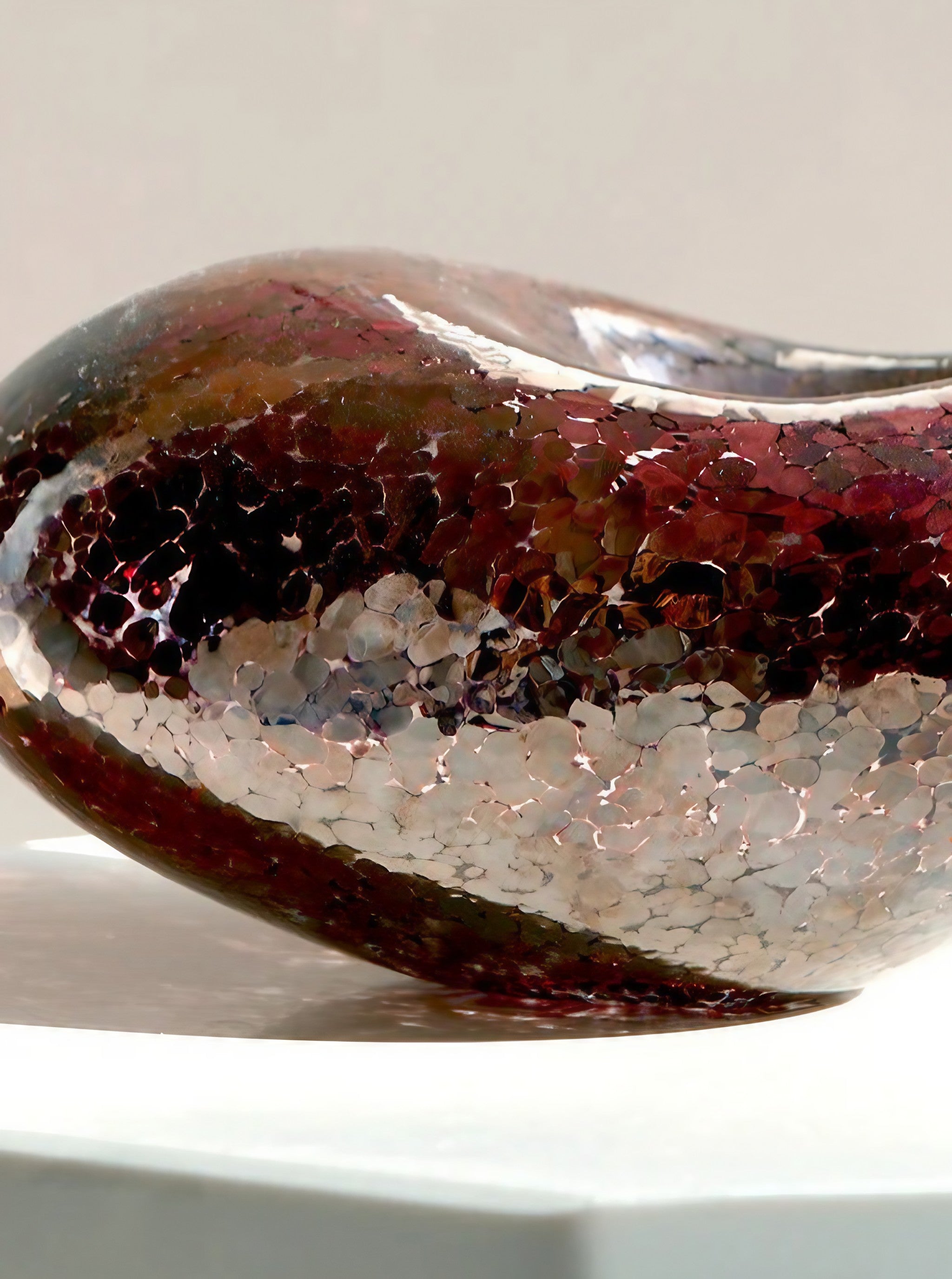 A detailed close-up of the Vanderohe Curio's Lucky Bean in Copper shows off its contemporary, glossy sculpture with a smooth, curved design. The surface is textured and adorned with a mosaic pattern of reflective red, brown, and silver tiles. Its exquisite craftsmanship makes it an ideal piece for home decor, sitting elegantly on a white surface against a neutral background.